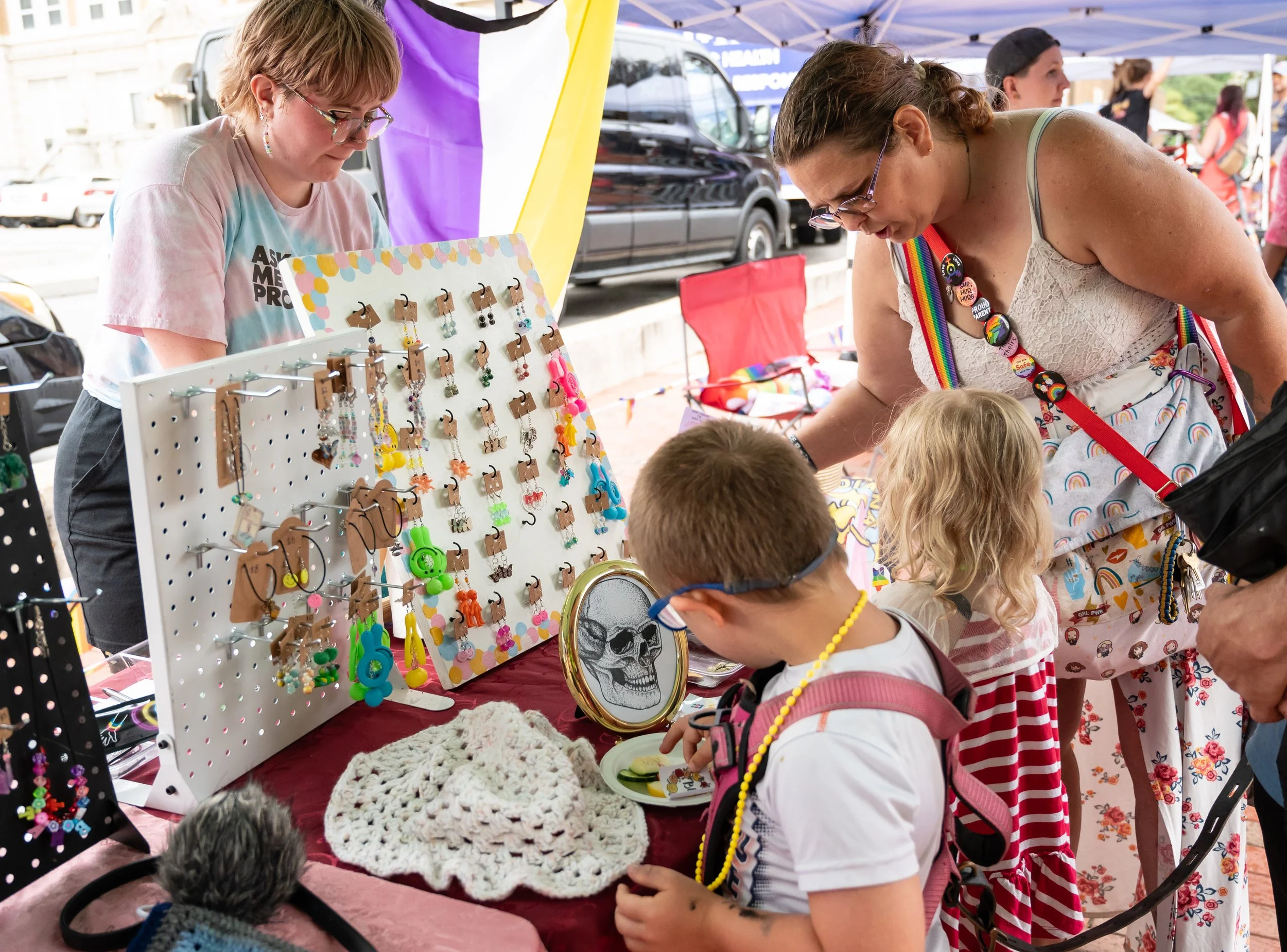 People browsing a jewelry booth at an outdoor event, with jewelry and accessories displayed on a white pegboard and table.