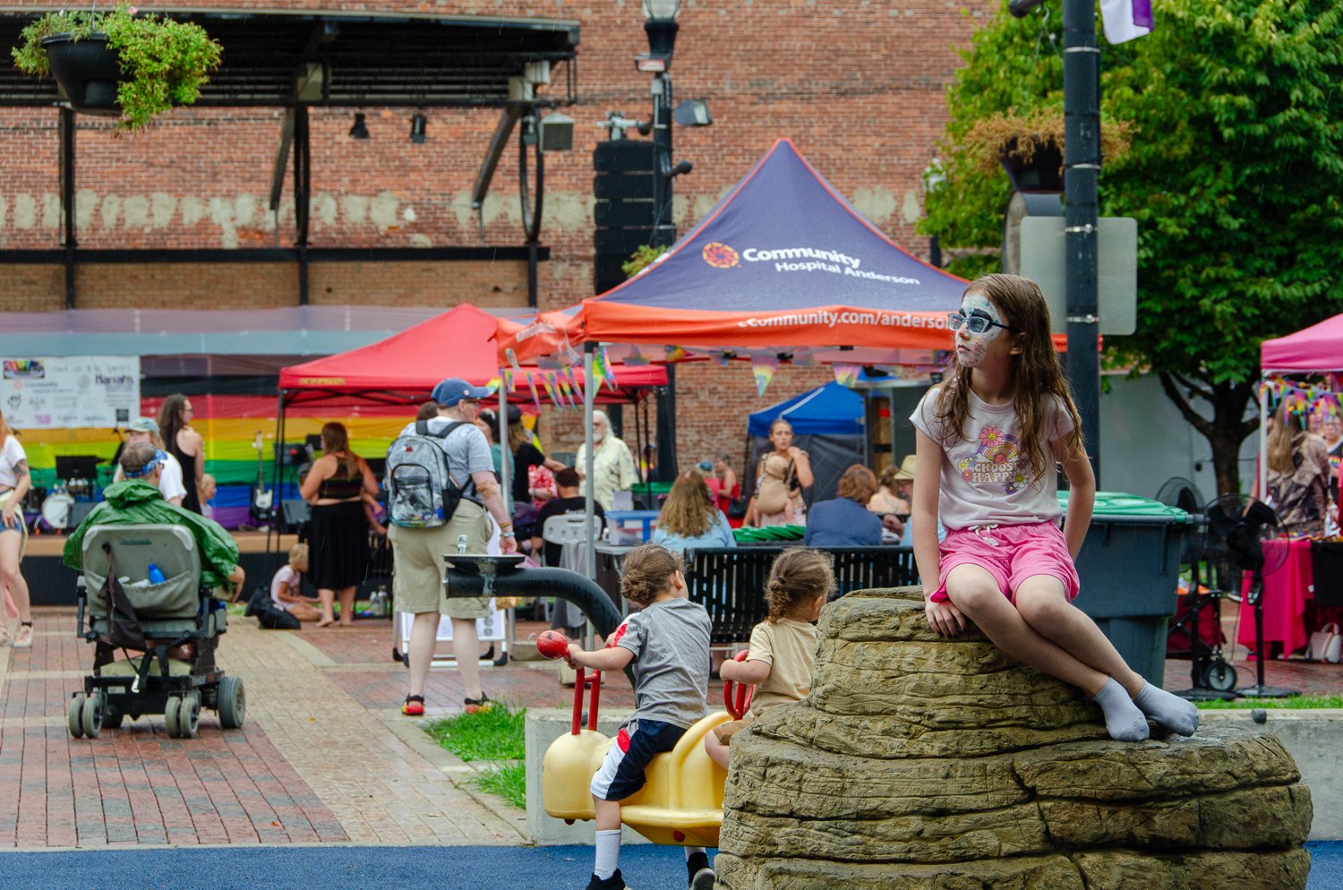 Children playing on a rock and a toy motorcycle, with a community event and rainbow flag tents in the background, and a girl with face paint sitting on a rock.