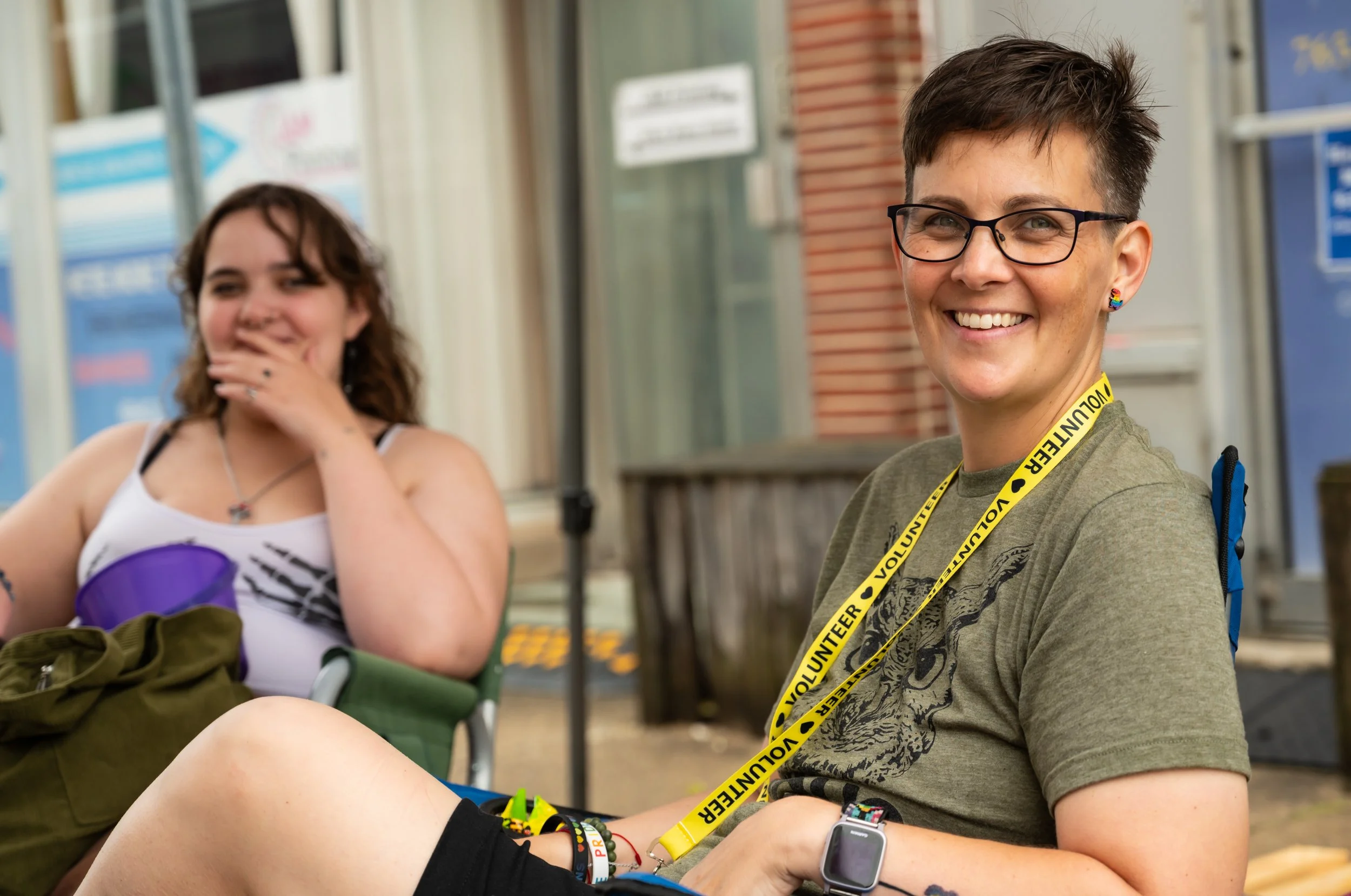 Two smiling women seated outdoors, one wearing glasses and a volunteer lanyard, with the other woman partially visible in the background.