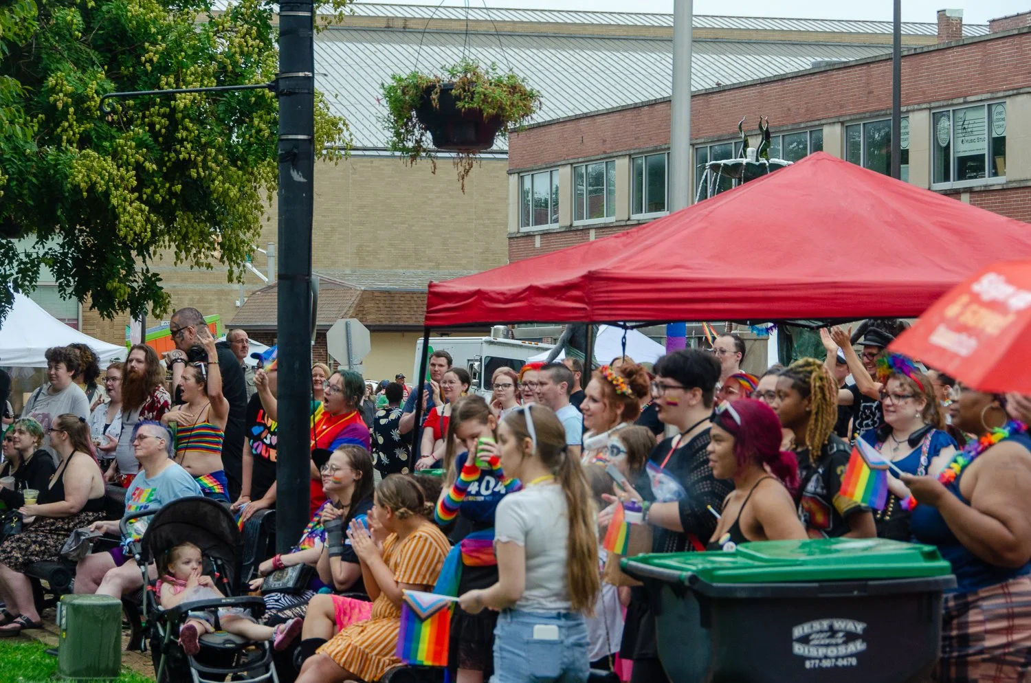 A crowd of people at Anderson Pride, many dressed in rainbow-colored clothing and accessories, with some holding rainbow flags, under a red canopy tent.