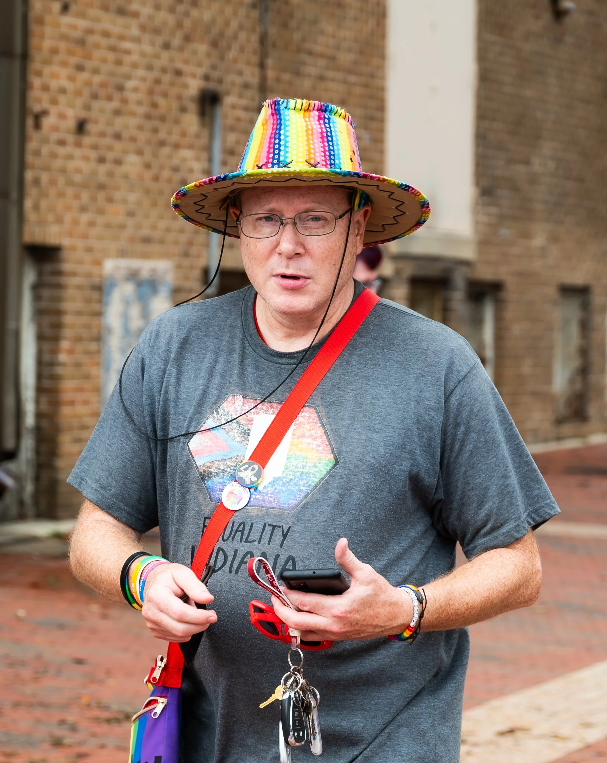 A man wearing a rainbow-colored straw hat with rainbow wristbands, holding a phone, with keys and car remote attached to a red lanyard, standing outdoors on a brick sidewalk with brick buildings in the background.