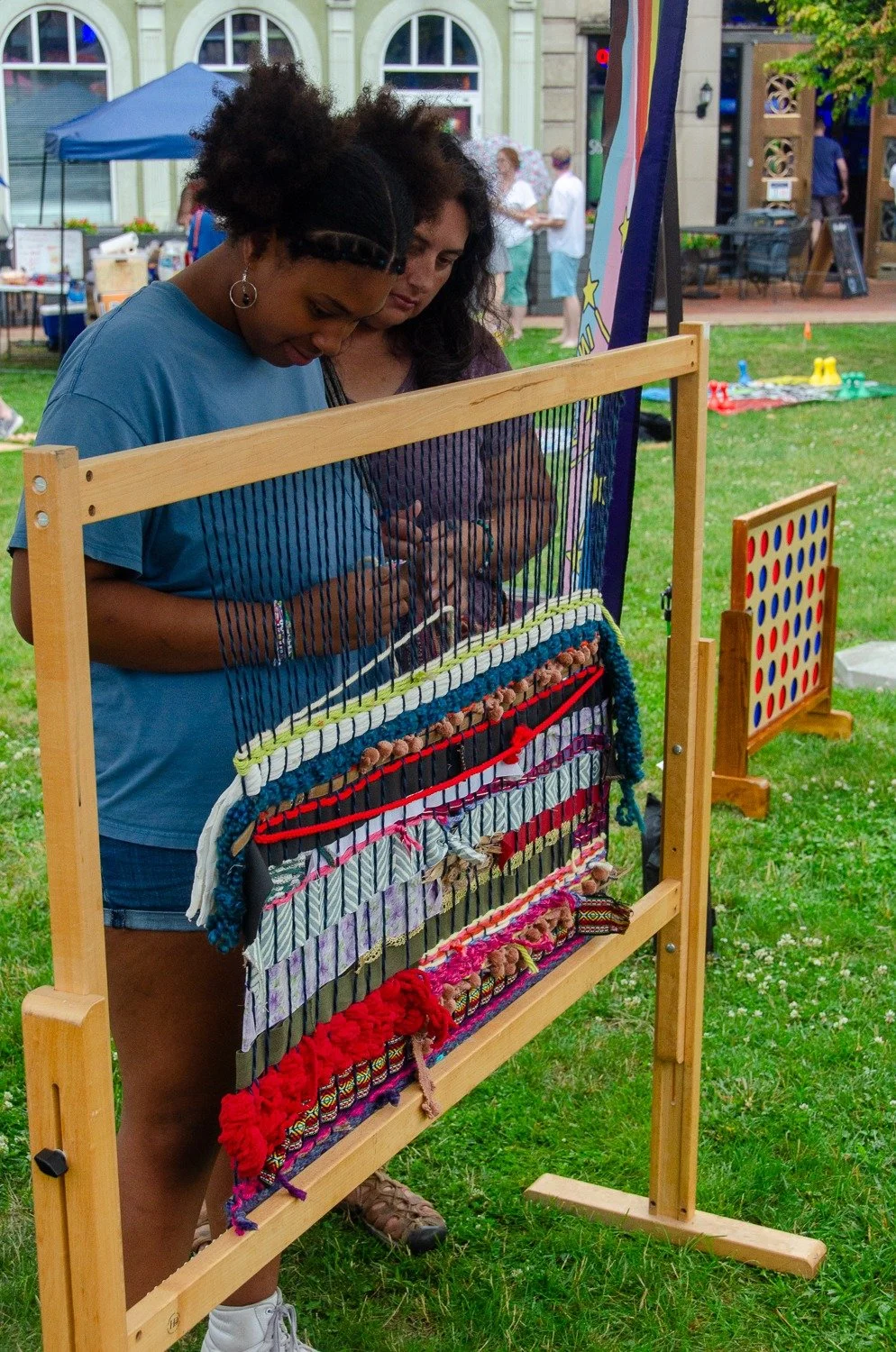 Two women are playing homemade giant connect four at an outdoor event, with green grass and booths in the background.