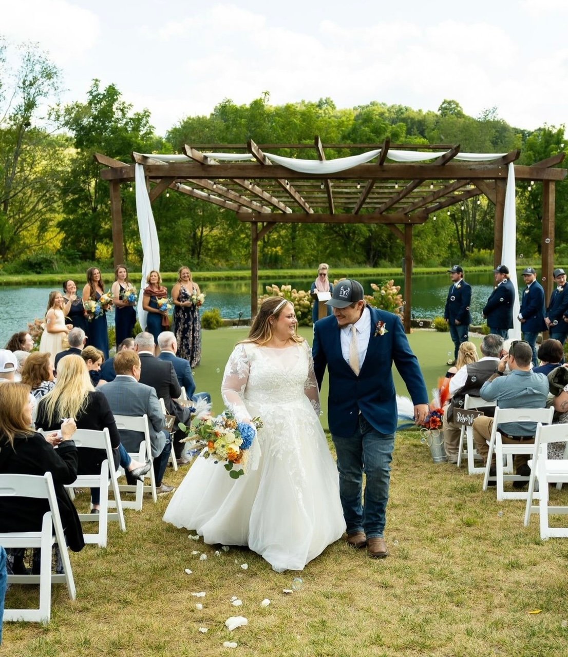 Bride and groom walking down the aisle during an outdoor wedding ceremony by a lake, with guests seated on white chairs and bridesmaids and groomsmen in the background.