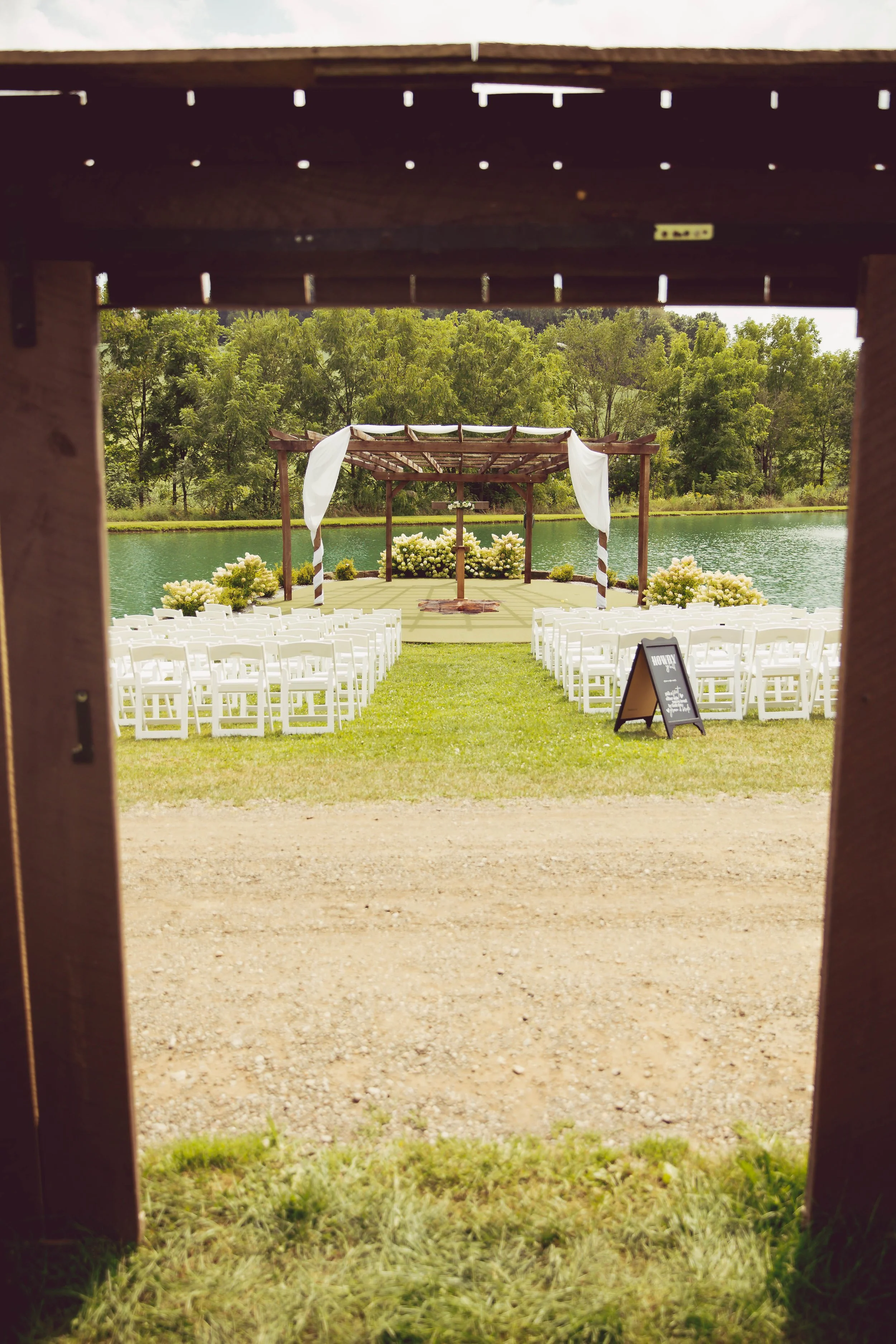 Outdoor wedding ceremony setup by a lake with white chairs and a decorated wooden archway.