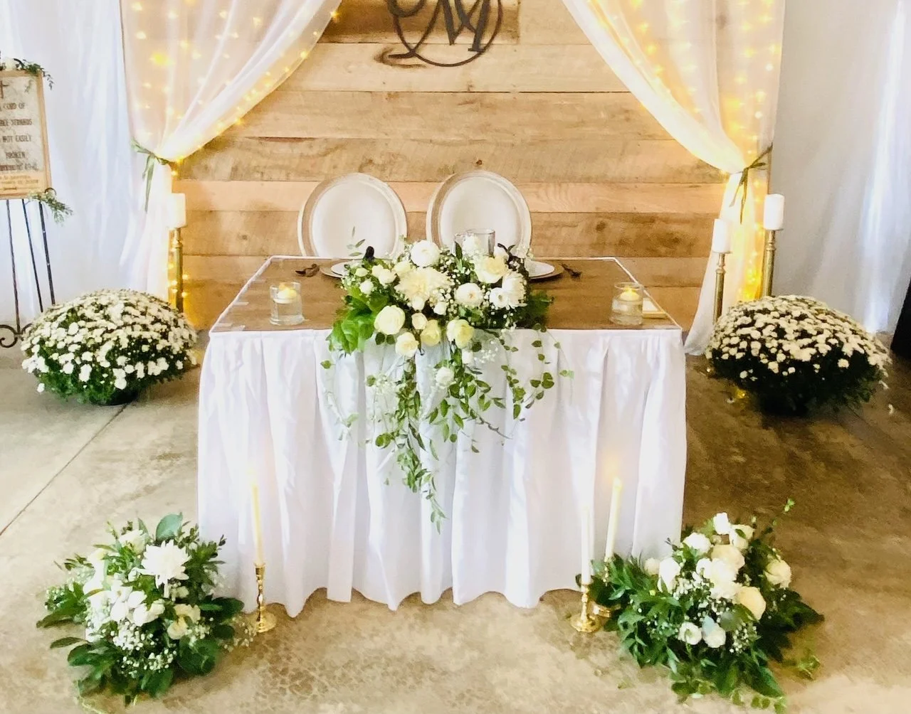 Wedding or event reception table with white drapery and fairy lights, decorated with white roses and green foliage, candlesticks, and candles for an elegant celebration.