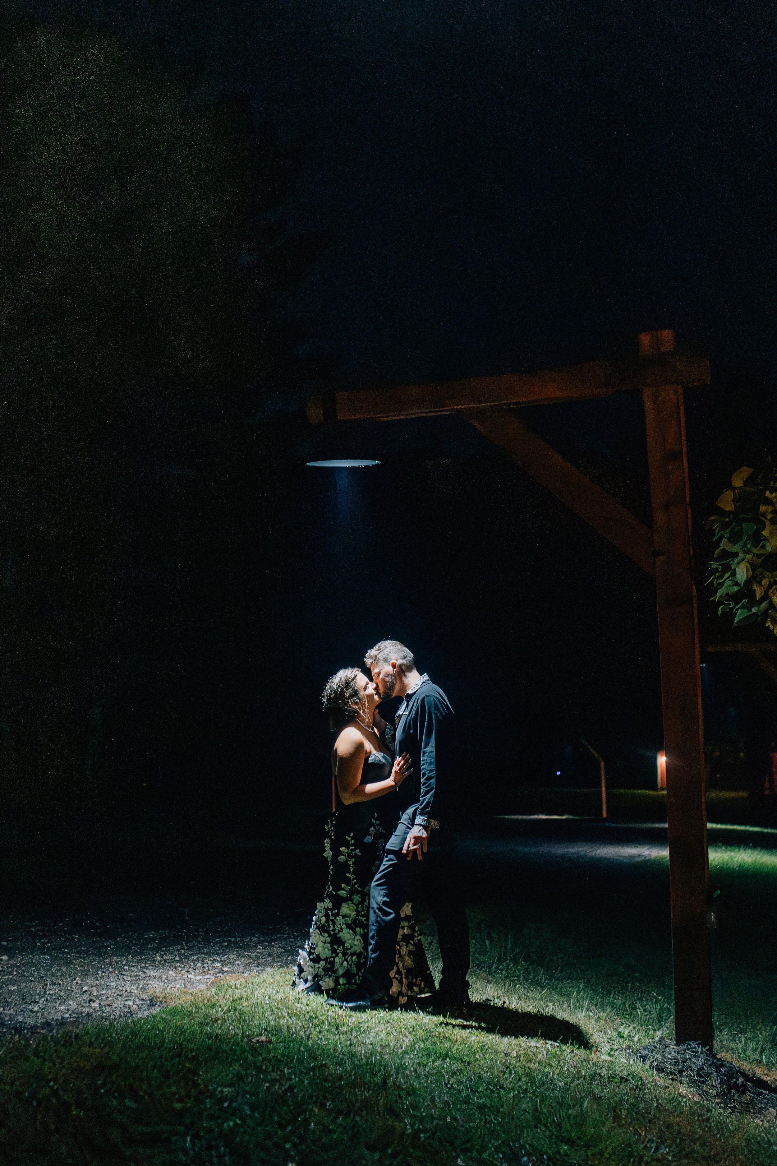 A couple sharing a romantic kiss at night illuminated by a spotlight, standing under a wooden arch in an outdoor setting.