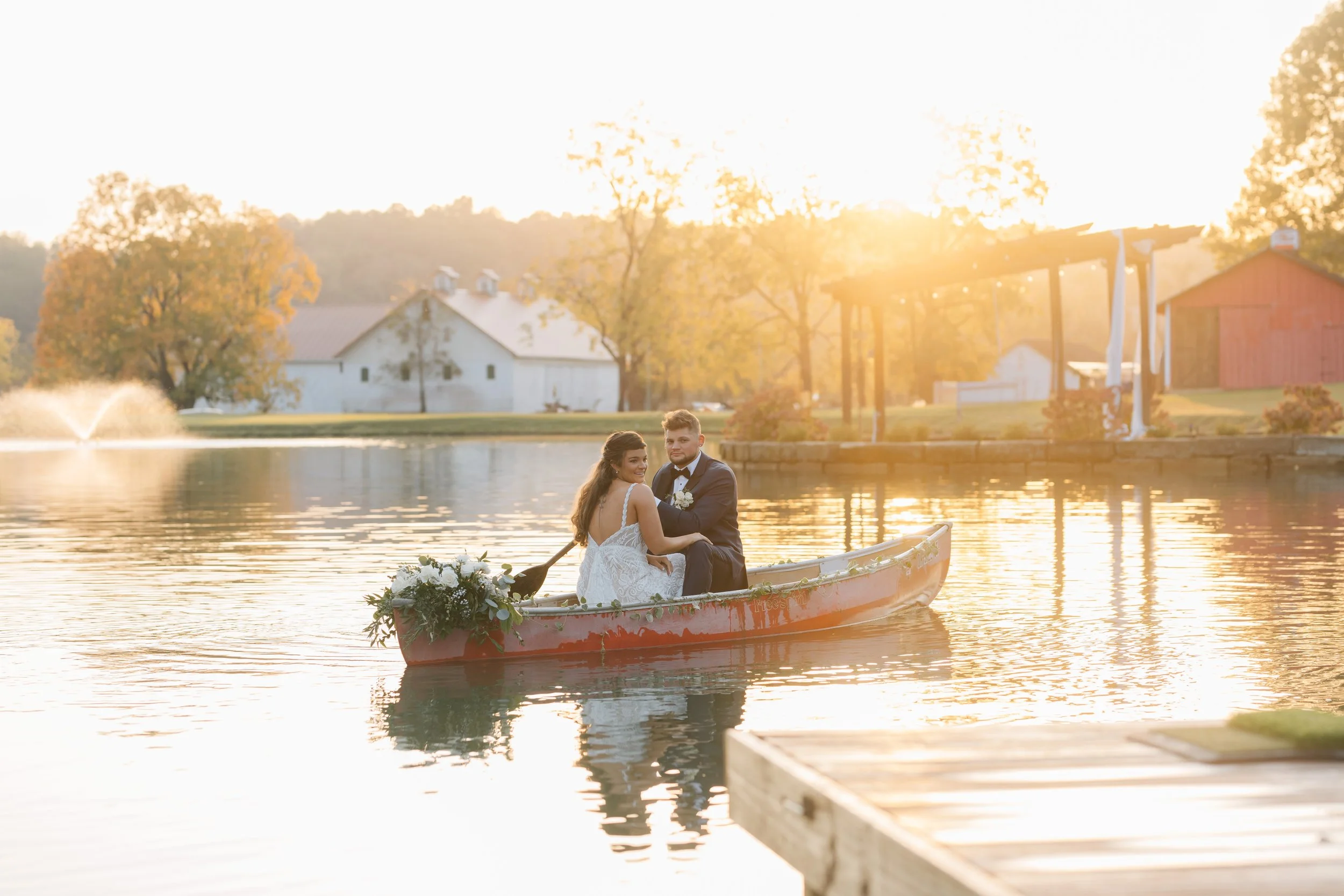 A bride and groom sitting in a boat decorated with flowers, on a lake during sunset, with farm buildings and trees in the background.