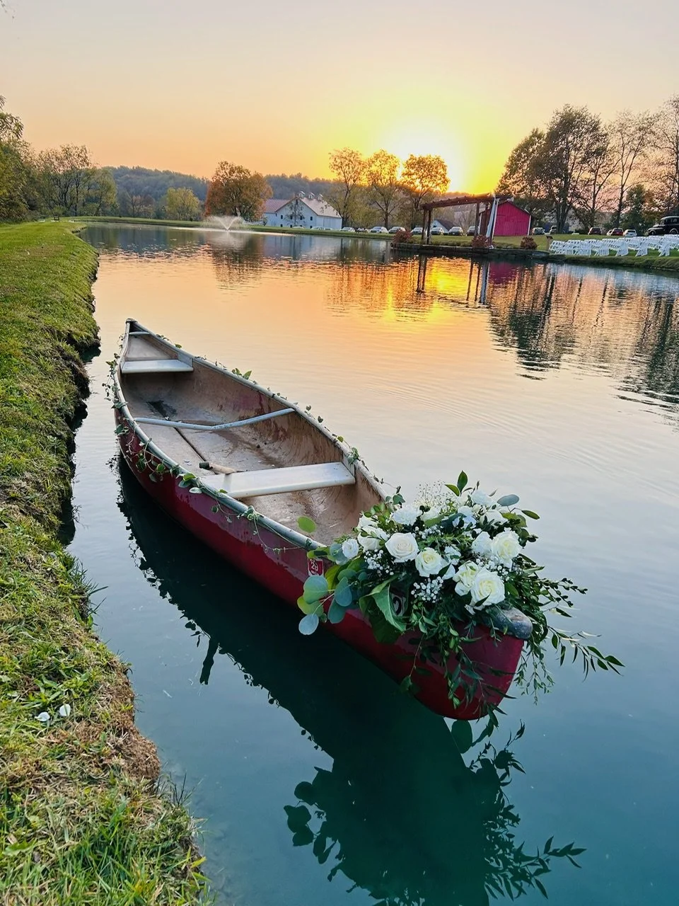 A red canoe decorated with white roses and greenery is floating on a calm river at sunset, with trees and a small building in the background.