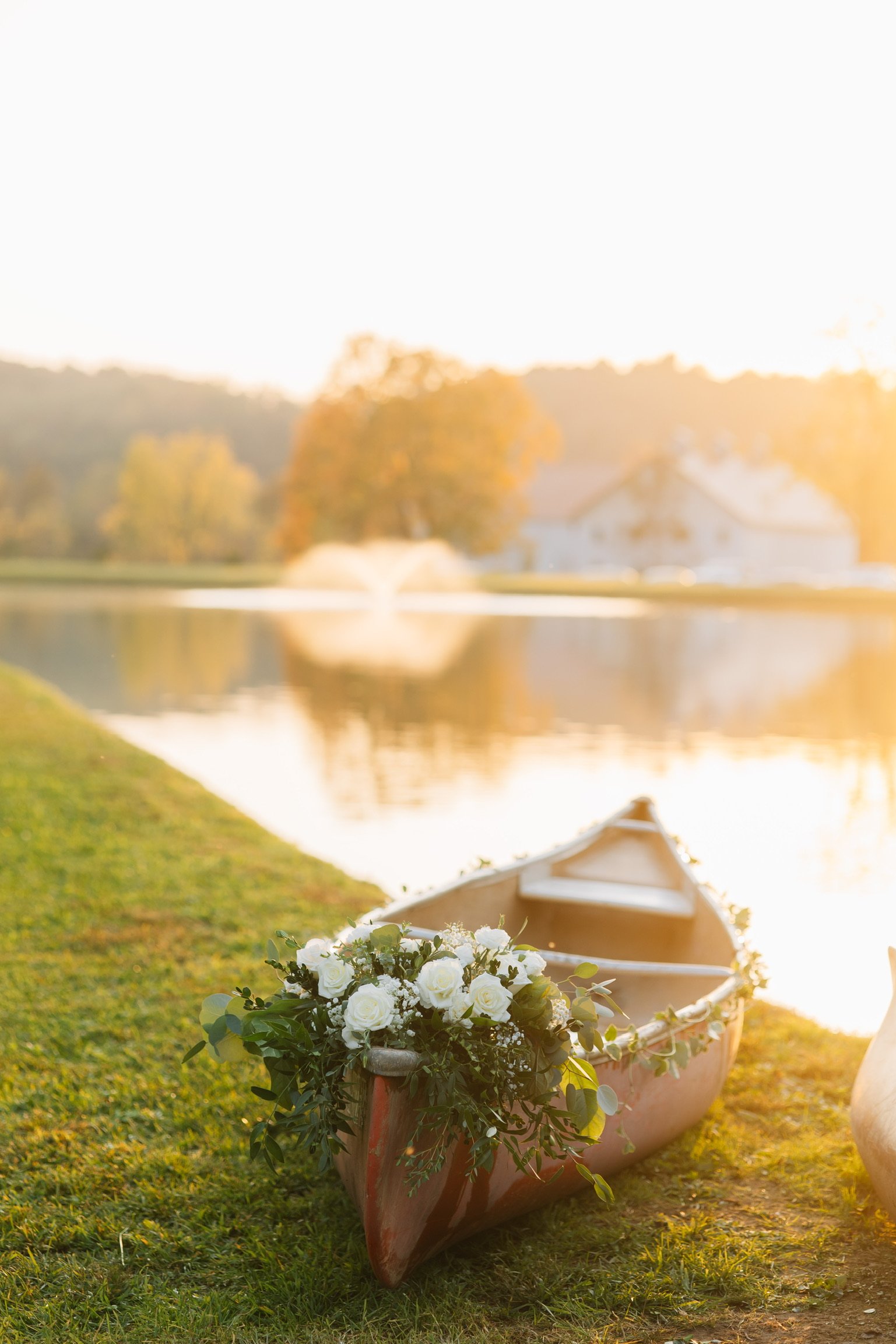 A canoe decorated with white roses and greenery rests on grass beside a lake during sunset, with a fountain, trees, and a house reflected in the water.