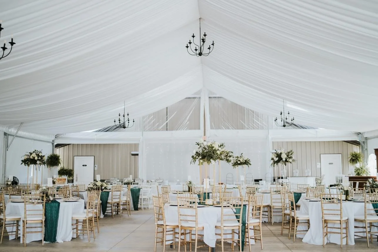 Elegant white event tent decorated with chandeliers, tables with white tablecloths, gold chairs, and floral centerpieces for a celebration or wedding reception.