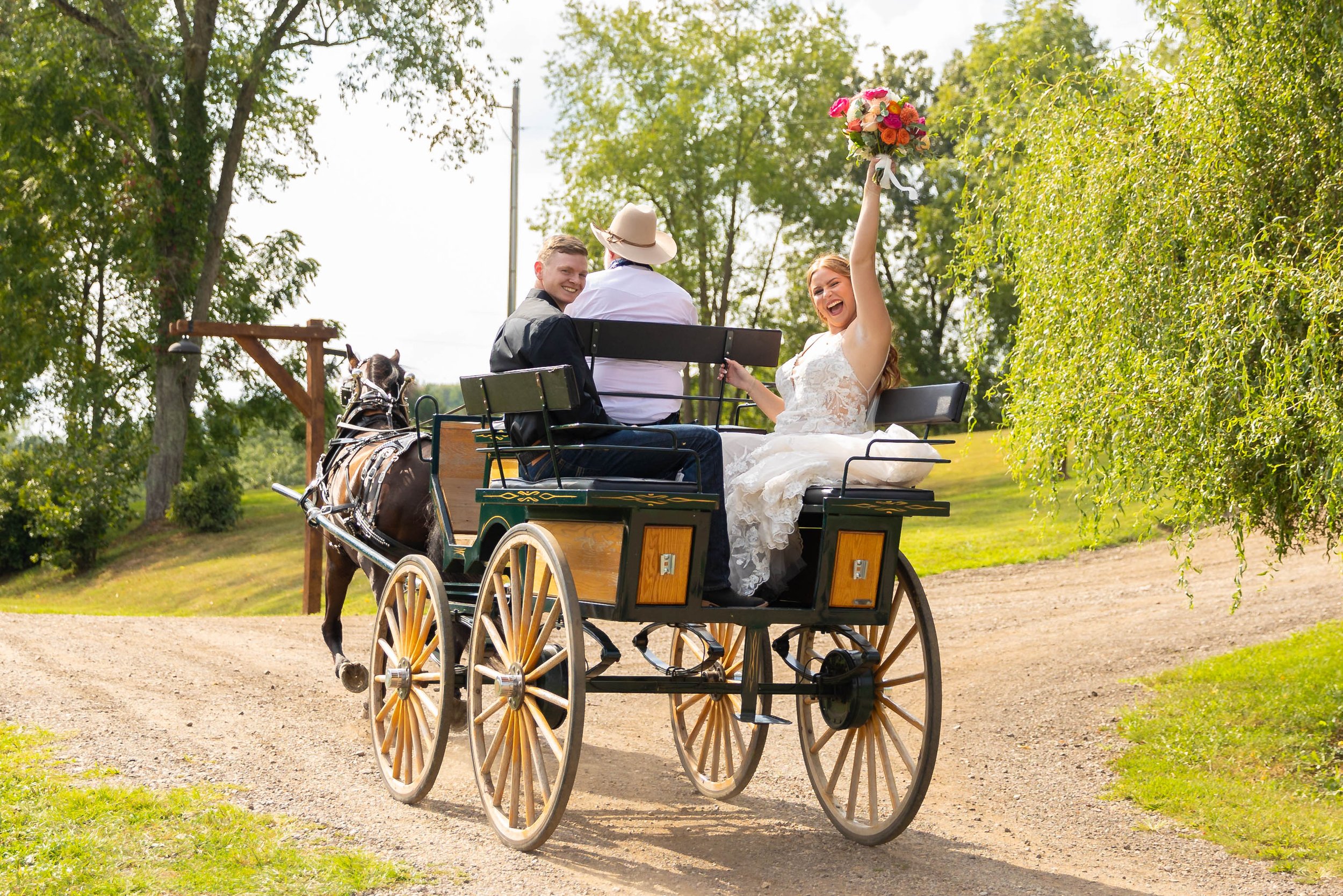 A bride in a wedding dress sitting in a horse-drawn carriage, holding a bouquet of flowers in the air, smiling. Two people are sitting beside her, one with a straw hat, on a dirt path with green trees in the background.