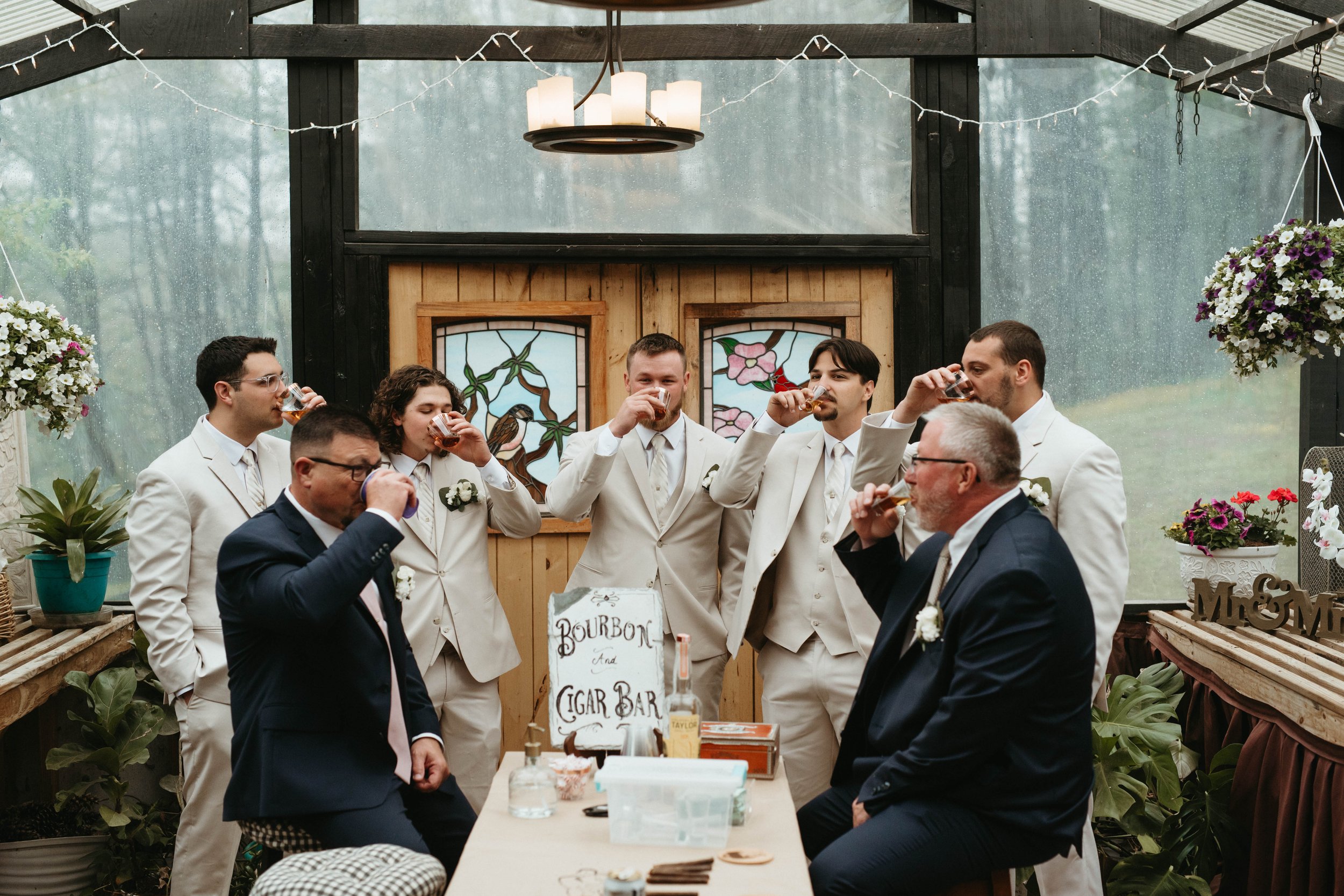 A group of people at a wedding celebration taking a shot together inside a rustic, greenhouse-style venue surrounded by plants and hanging flower baskets.