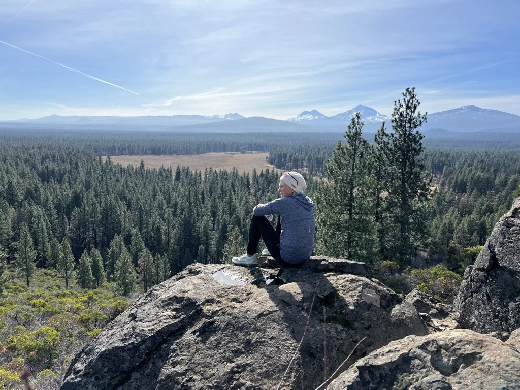 Woman sitting quietly on a rock overlooking a valley of trees and mountains, reflecting and finding peace in nature.