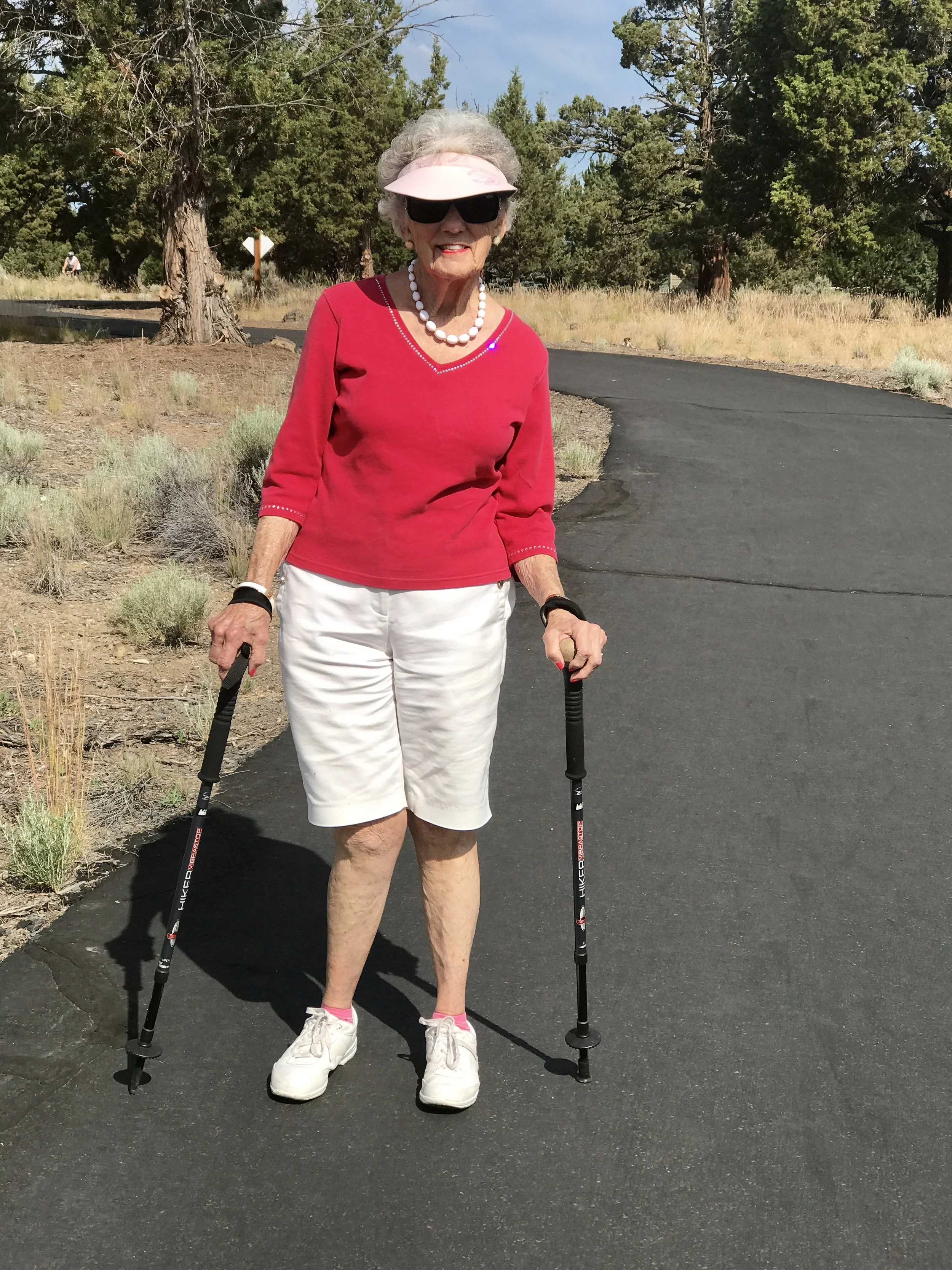 Active older woman walking with trekking poles on a paved trail, demonstrating balance, strength, and healthy movement in aging.