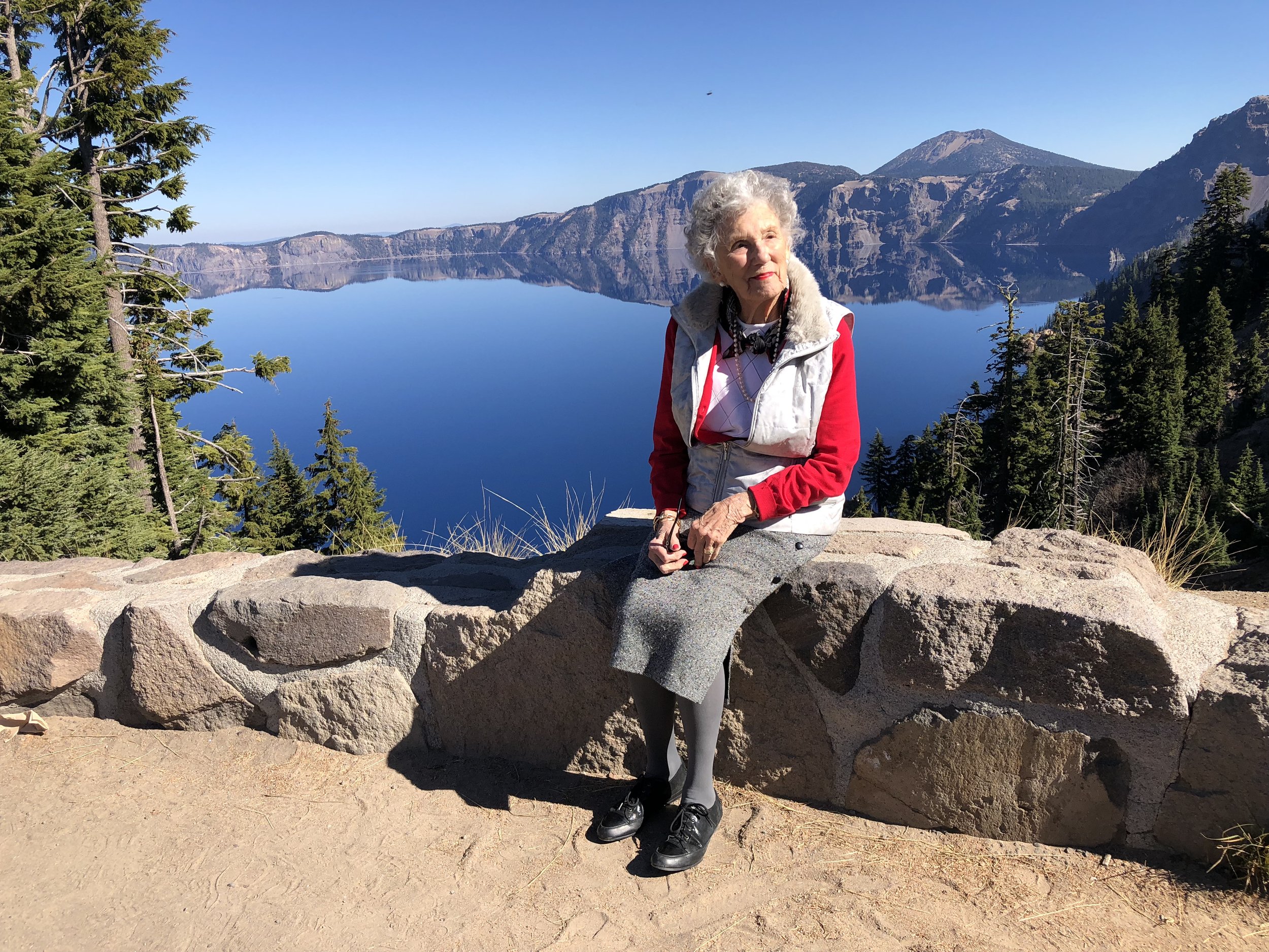 Elderly woman sitting at Crater Lake with the calm blue water in the background, enjoying nature and outdoor adventure in her later years.