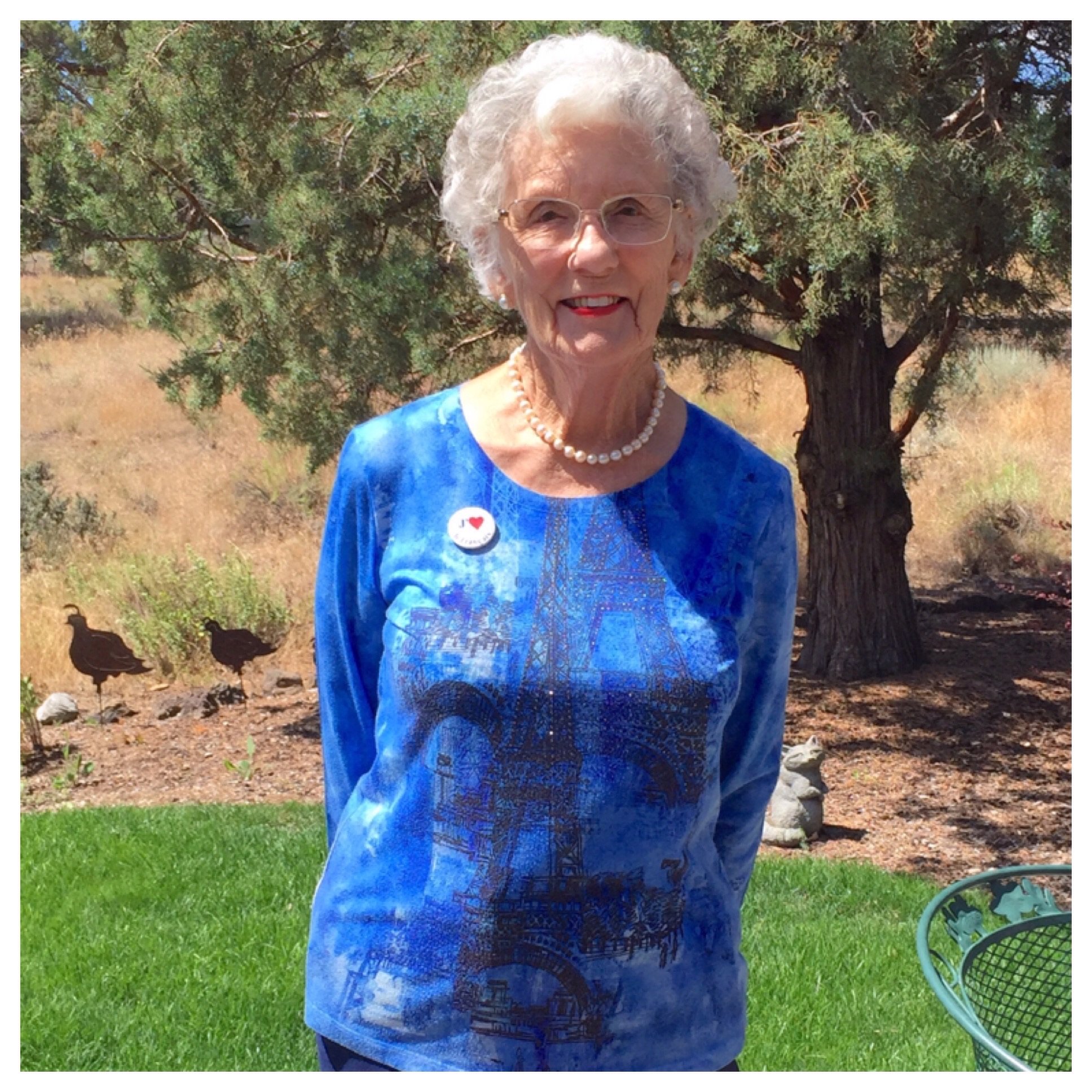 Smiling elderly woman standing outdoors in a blue top and pearls, embodying joy, beauty, and graceful aging.