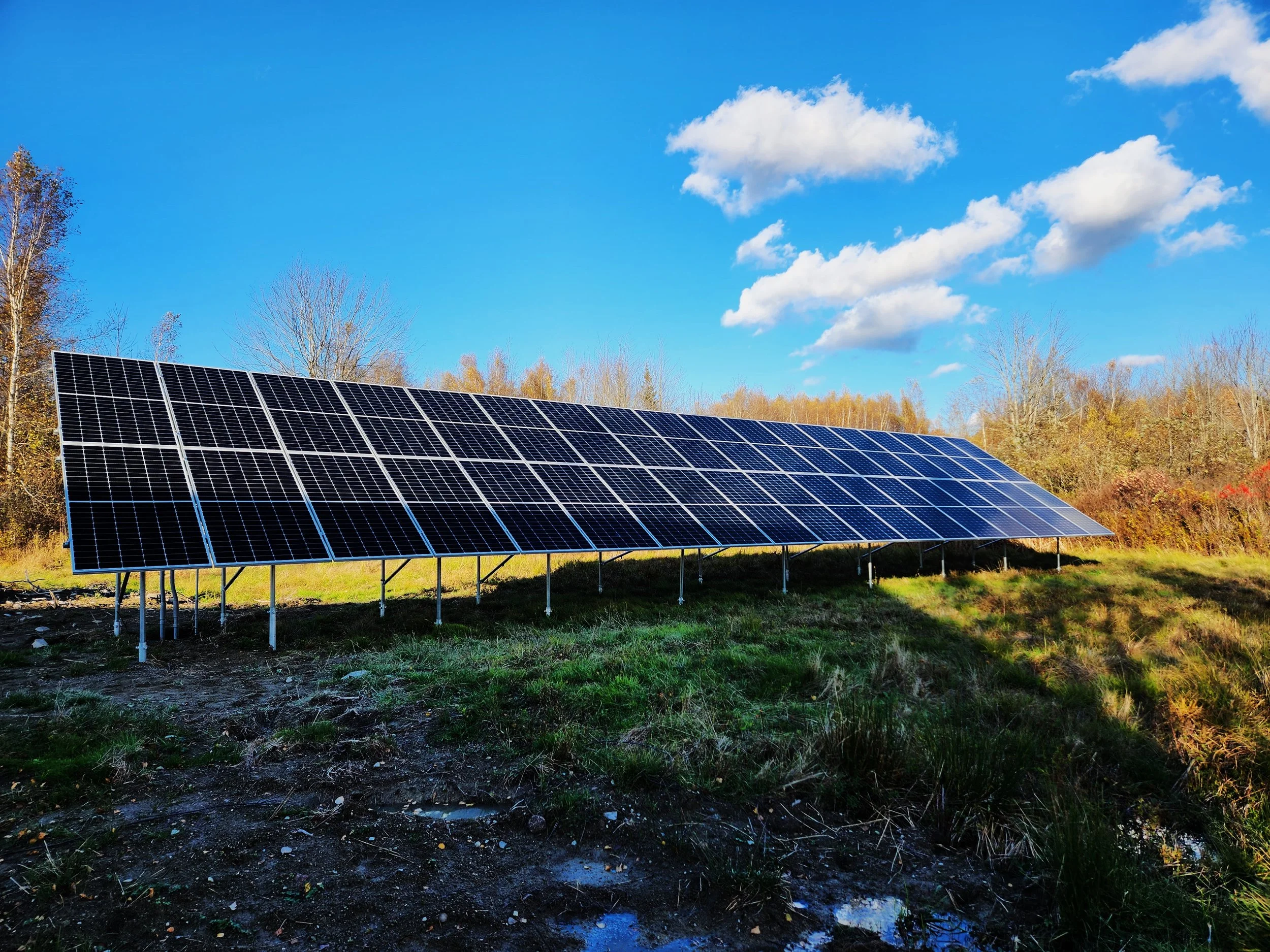 Solar panels installed in a grassy area with trees and a blue sky with clouds in the background.