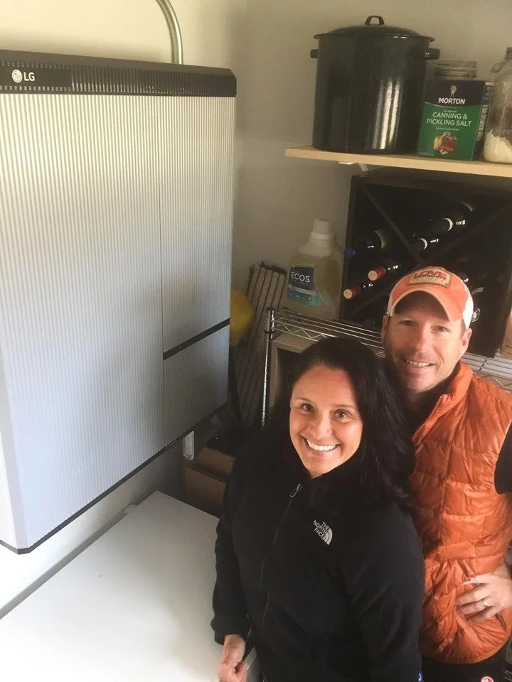 A smiling woman and man standing in a kitchen. The woman has black hair and is wearing a black North Face jacket. The man has a beard, is wearing a beige cap and an orange puffer vest. They are near a white counter and a wine rack with bottles. On the shelves, there are some containers including a large black pot, a box of pickling salt, and a spray bottle.