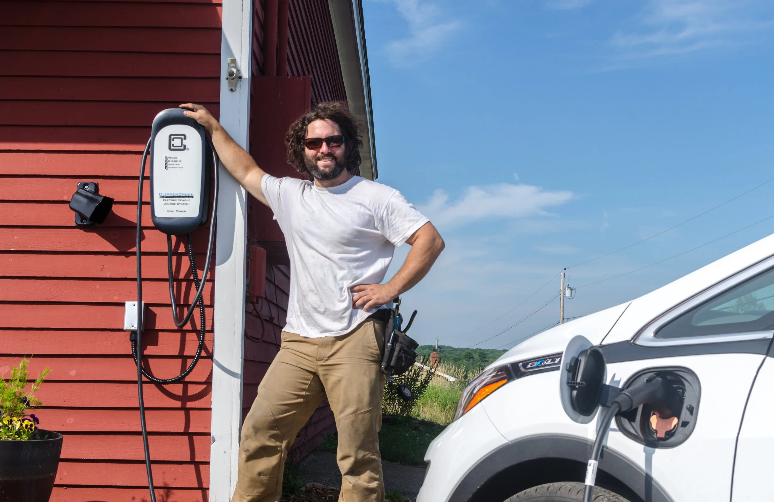 A man with curly hair, sunglasses, a white t-shirt, and khaki pants stands next to an electric vehicle charging station mounted on a red wooden building. The man has a tool belt and is smiling. A white electric car is plugged into the charging station, with a blue sky and power lines in the background.