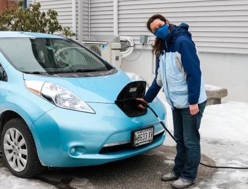 A woman charging an electric blue Nissan Leaf electric vehicle outdoors during winter, with snow on the ground, wearing a blue mask, jacket, and jeans.
