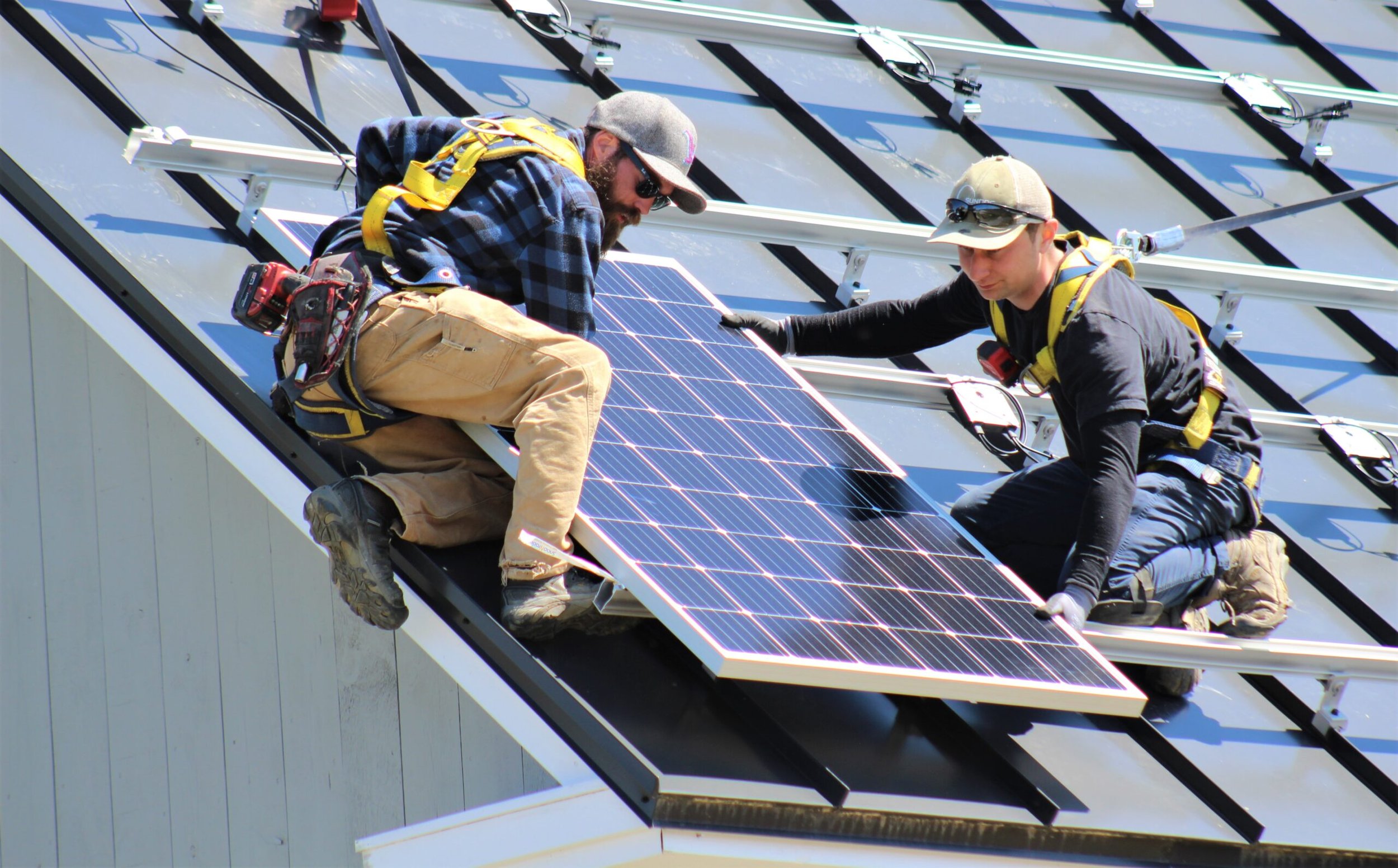 Two workers installing solar panels on a roof, wearing safety harnesses and using tools.