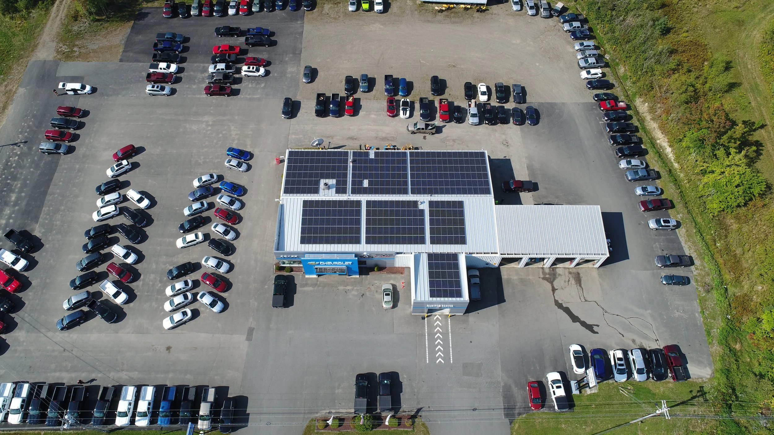 Aerial view of a car dealership lot filled with numerous cars of various colors, surrounding a building with solar panels on the roof, labeled 'Chevrolet'.