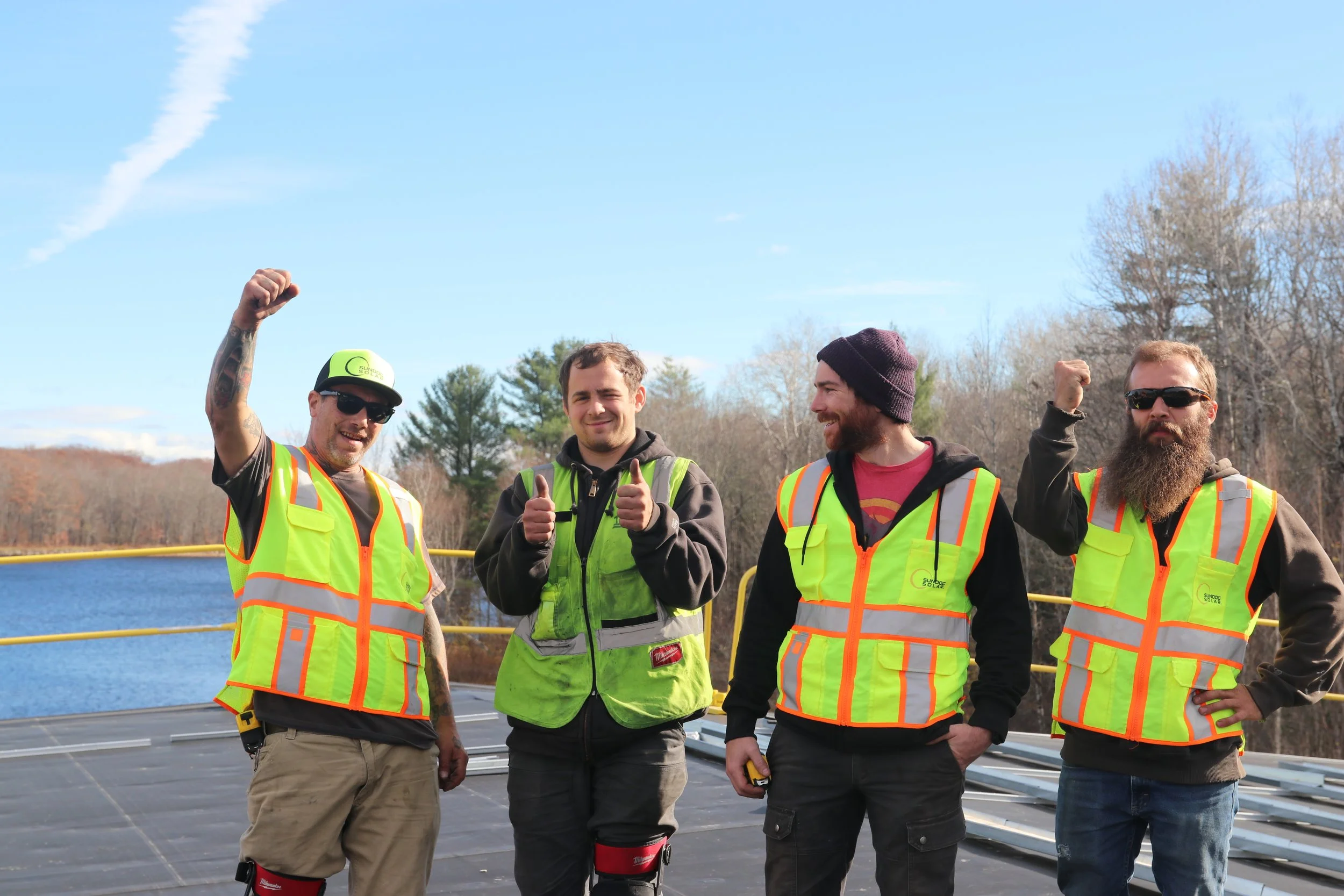 Four men standing outdoors on a sunny day, wearing high-visibility safety vests. One man on the far left is raising his arm in a fist, wearing sunglasses and a cap. The man second from the left is giving a thumbs-up, dressed in a black hoodie. The third man has a beard, is wearing a knit cap, and is smiling while looking at the man next to him. The man on the far right is also raising his fist and wearing sunglasses. Trees and a clear blue sky are visible in the background.