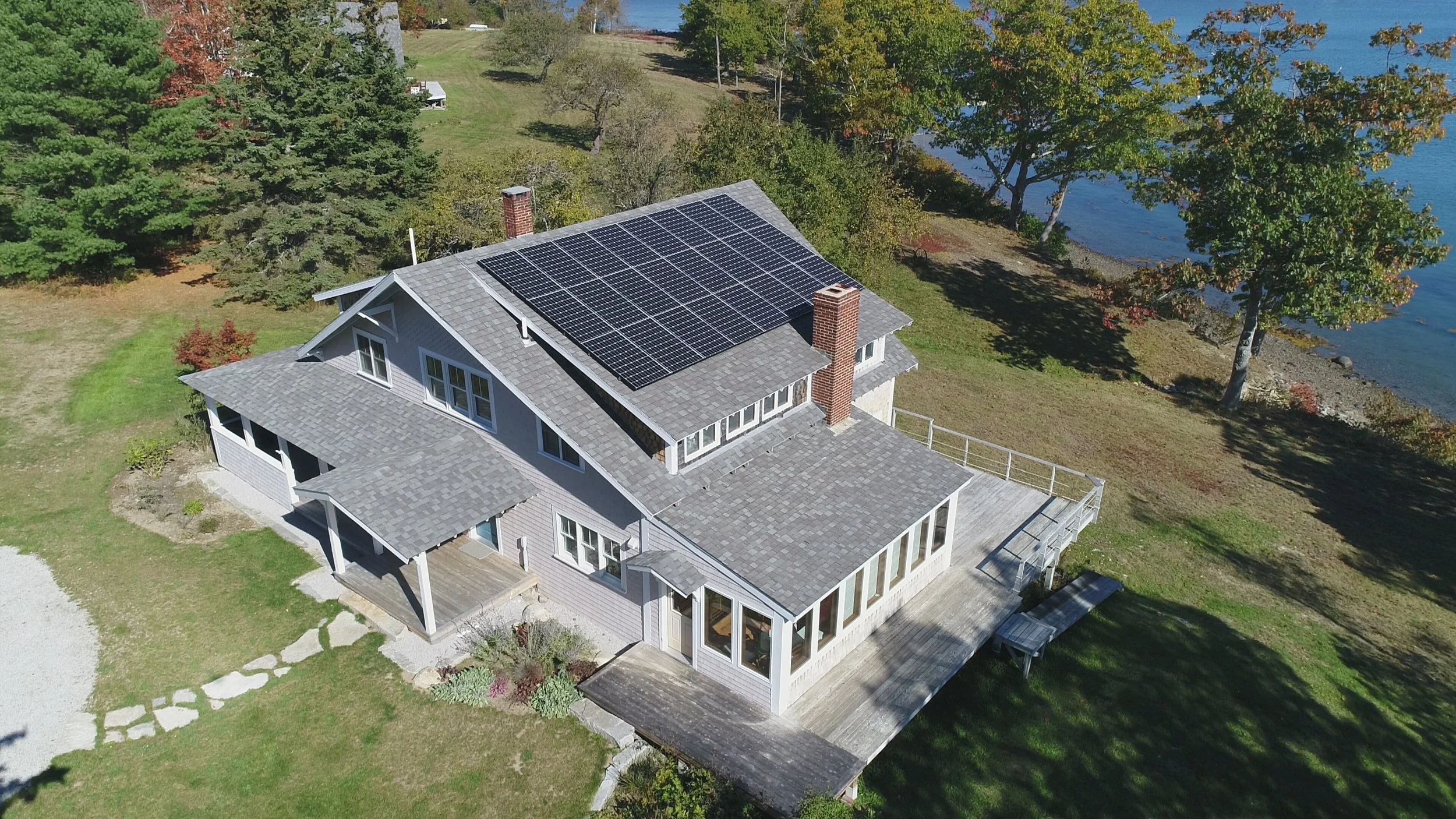 Aerial view of a house with solar panels on the roof, surrounded by trees and next to a body of water.