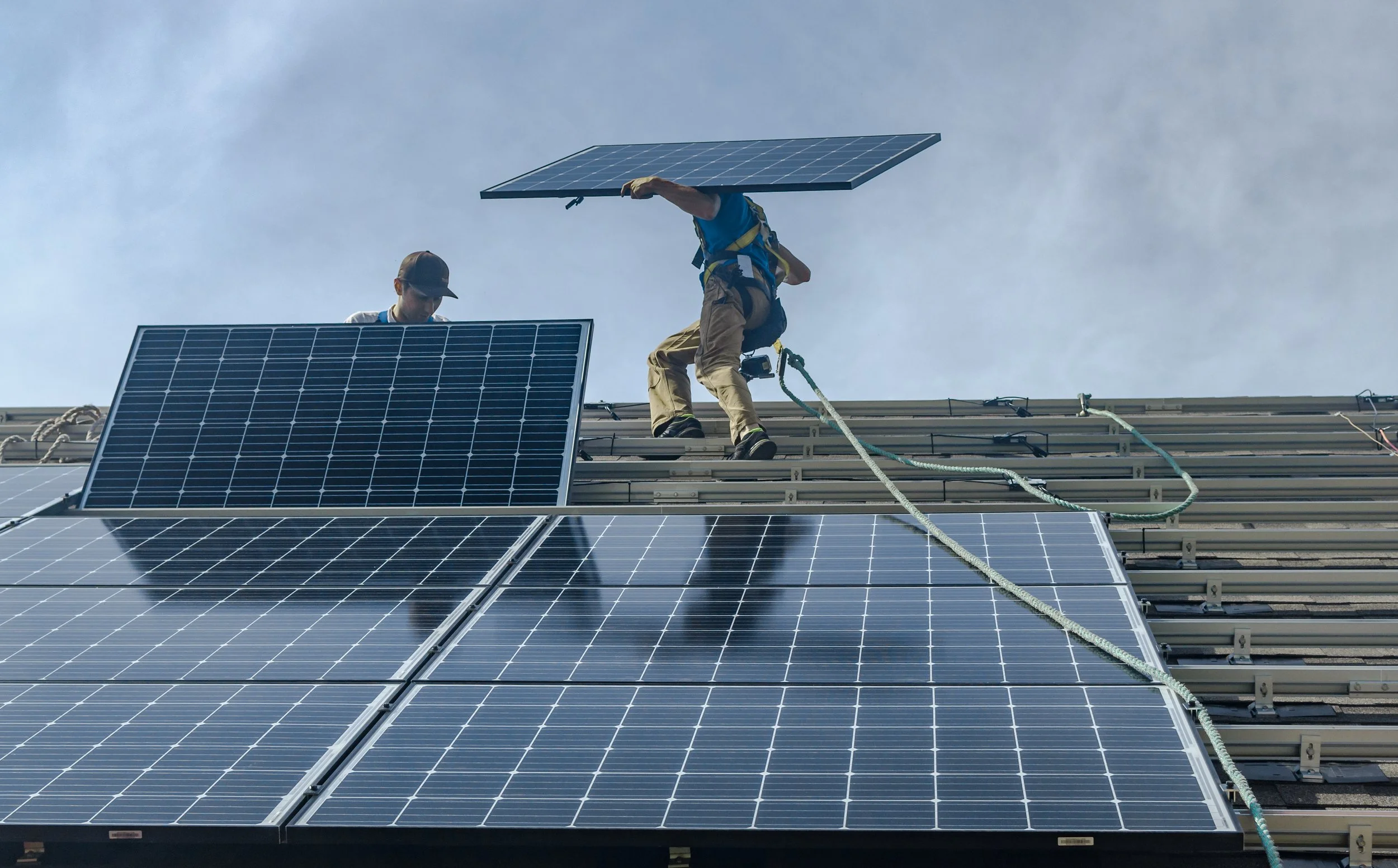 Two workers installing solar panels on a rooftop during daytime, one standing and the other crouching, with the sky in the background.