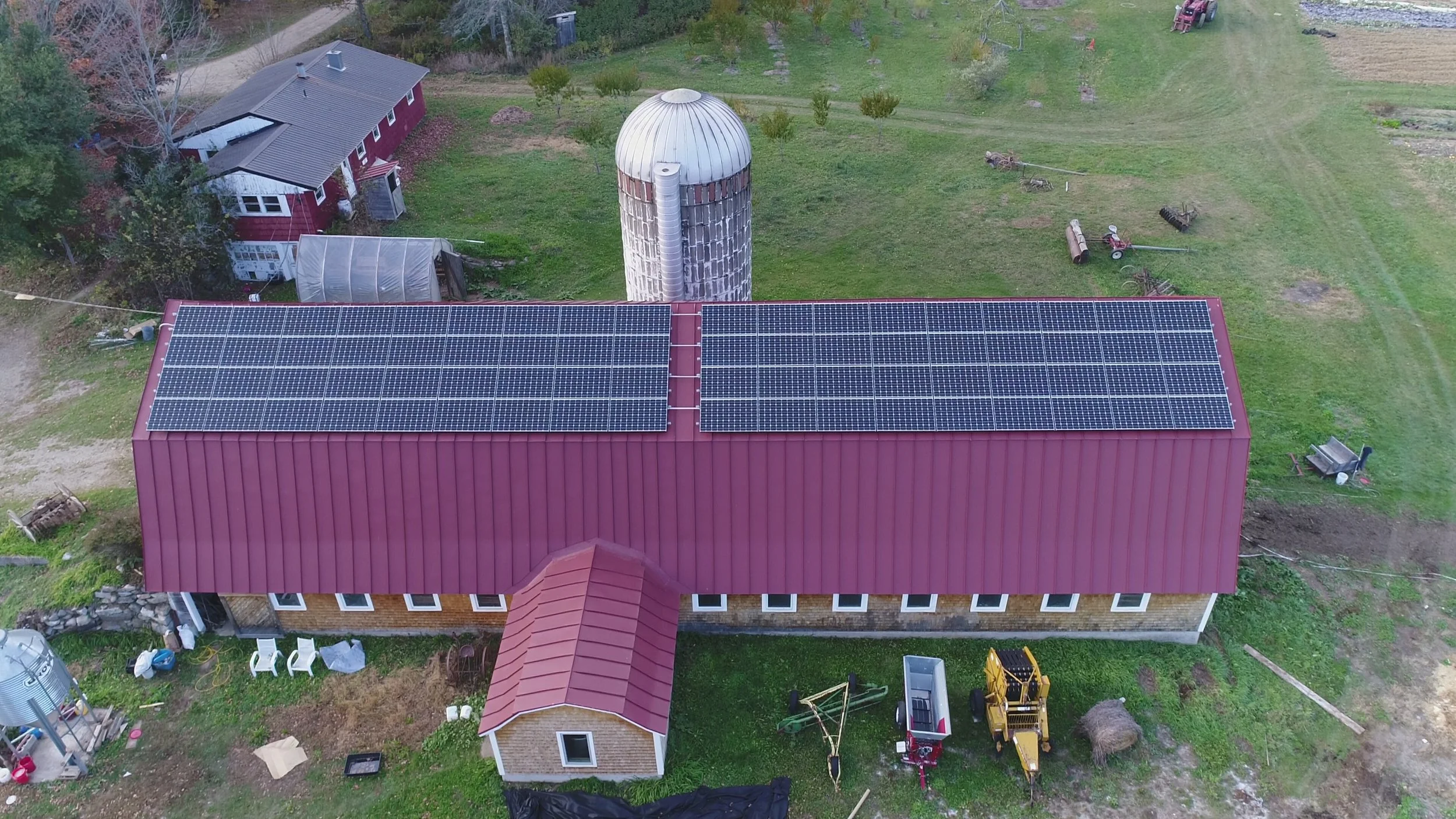 Aerial view of a farm building with a red metal roof covered with solar panels; in the background, a white grain silo, a small red house with a gray roof, and farmland with some equipment and trees.