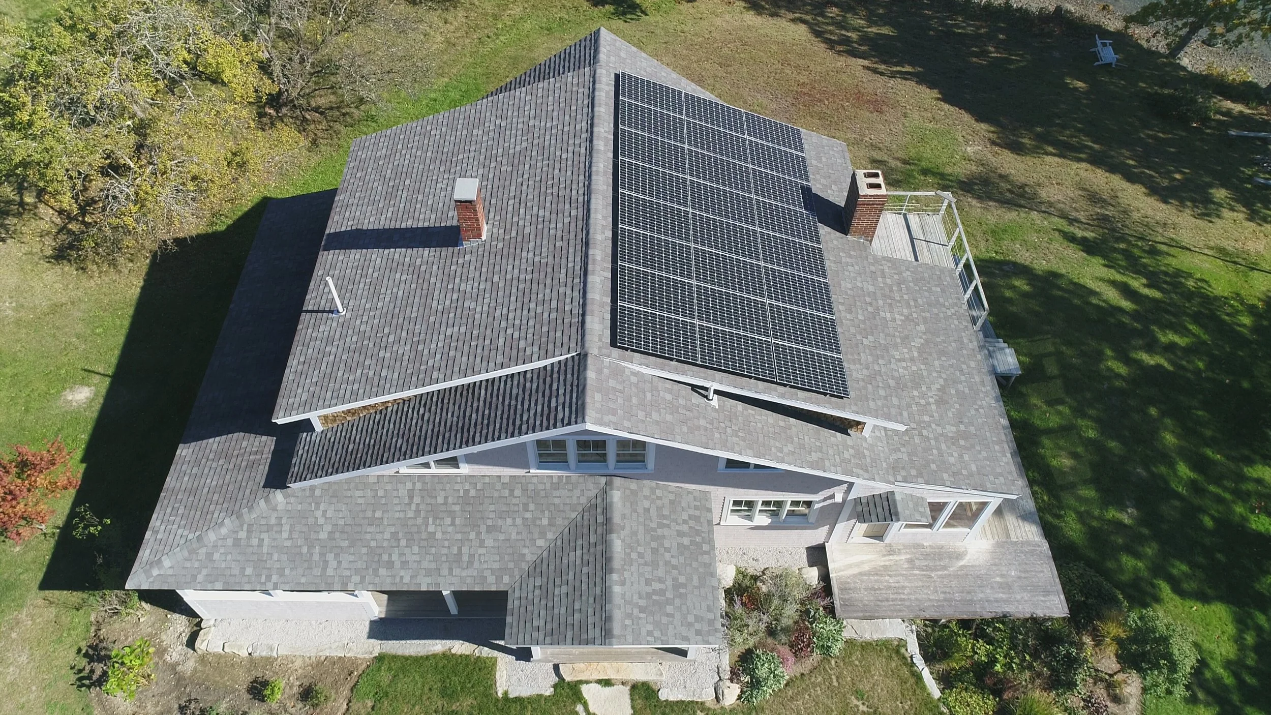 Aerial view of a house with solar panels on the roof, surrounded by grass, trees, and a backyard with a white chair.