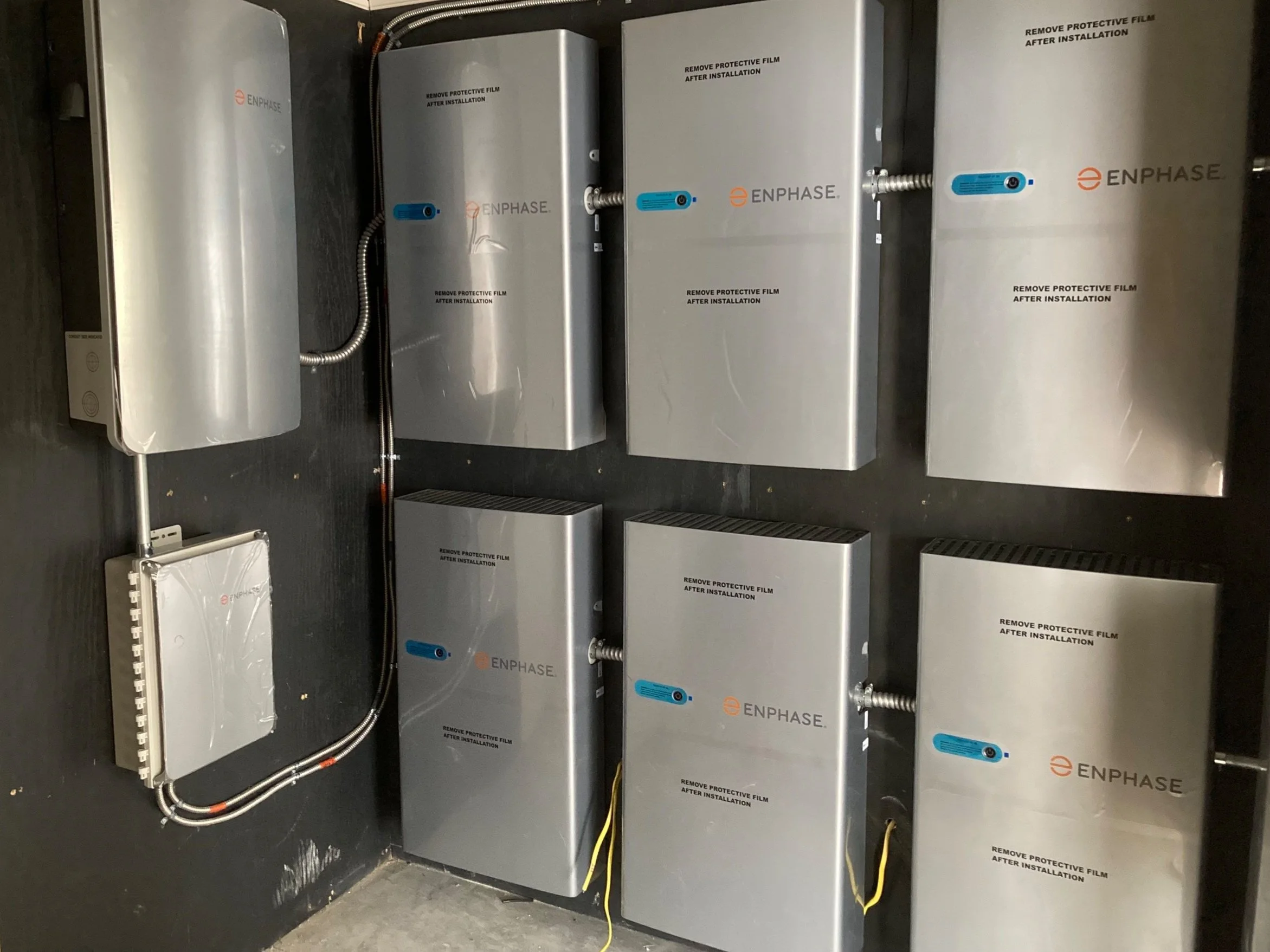 Multiple silver electrical enclosures mounted on a black wall with wiring cables connected. The enclosures are labeled with the brand 'ENPHASE' and have instructions to remove protective film after installation.