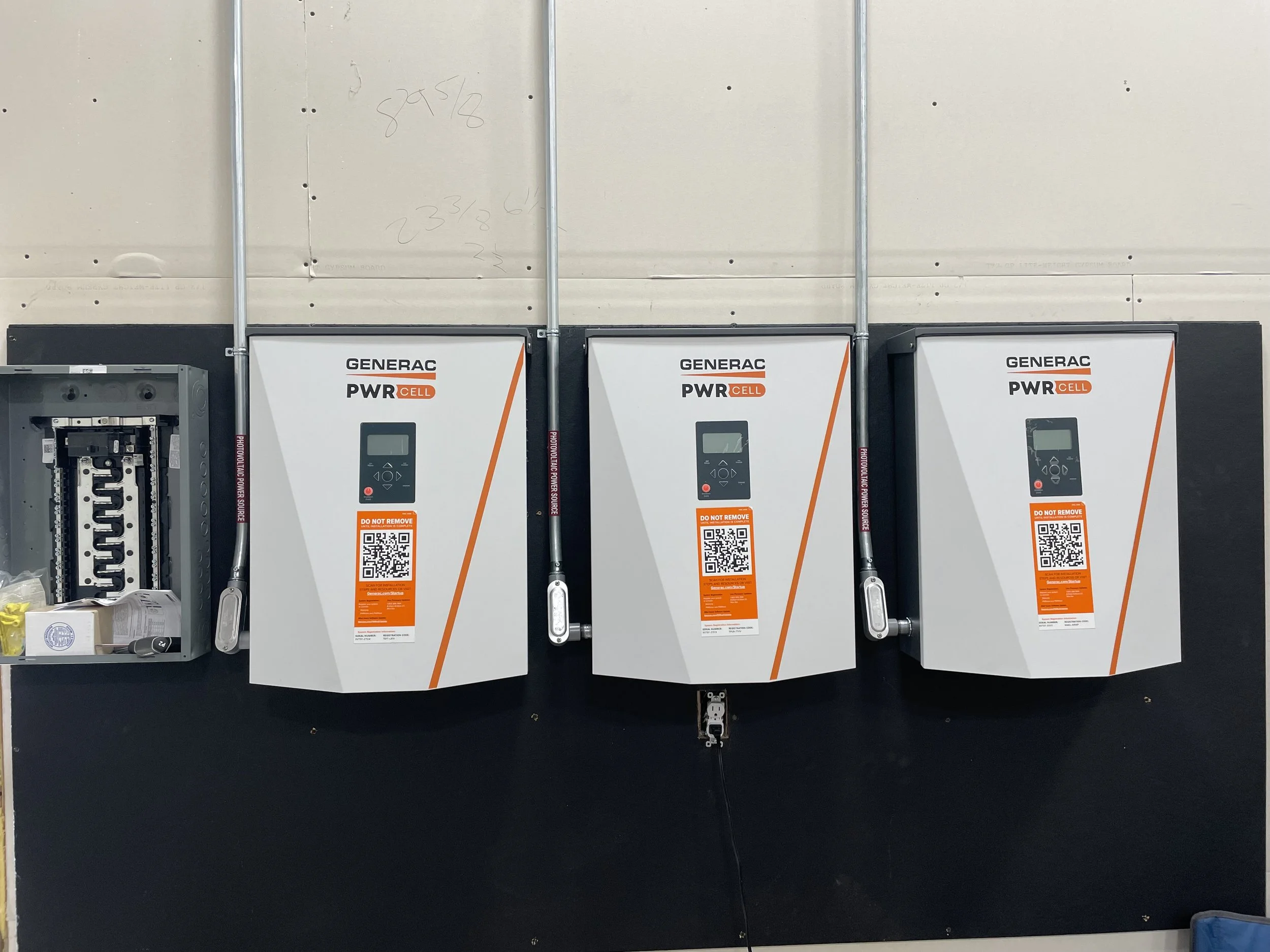 Three Generac PWRcell solar inverter units mounted on a wall in a technical or utility room. To the left, part of an electrical panel with wiring and components is visible. The units have digital displays and QR codes on orange labels.
