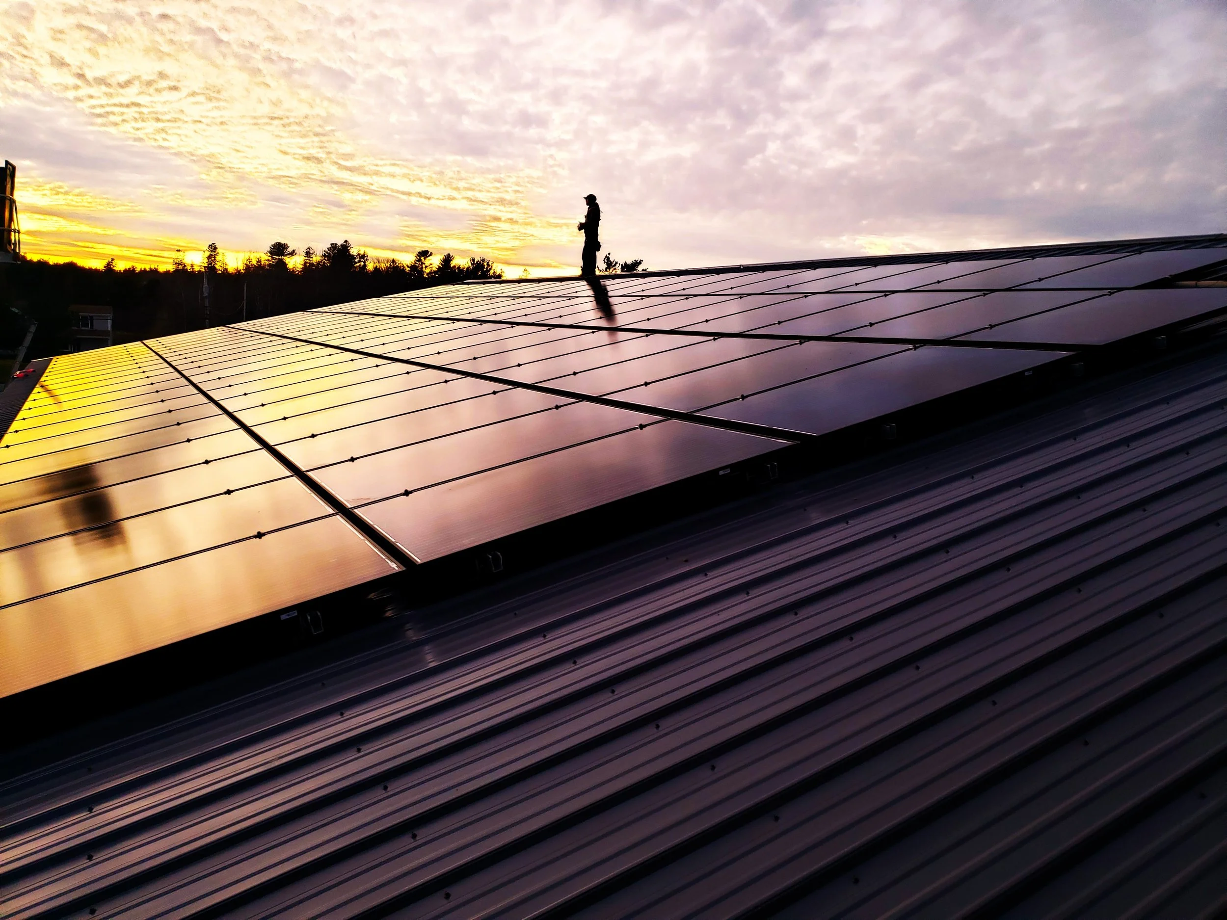 A person standing on a roof covered with solar panels during sunset, with a sky filled with clouds and trees in the background.