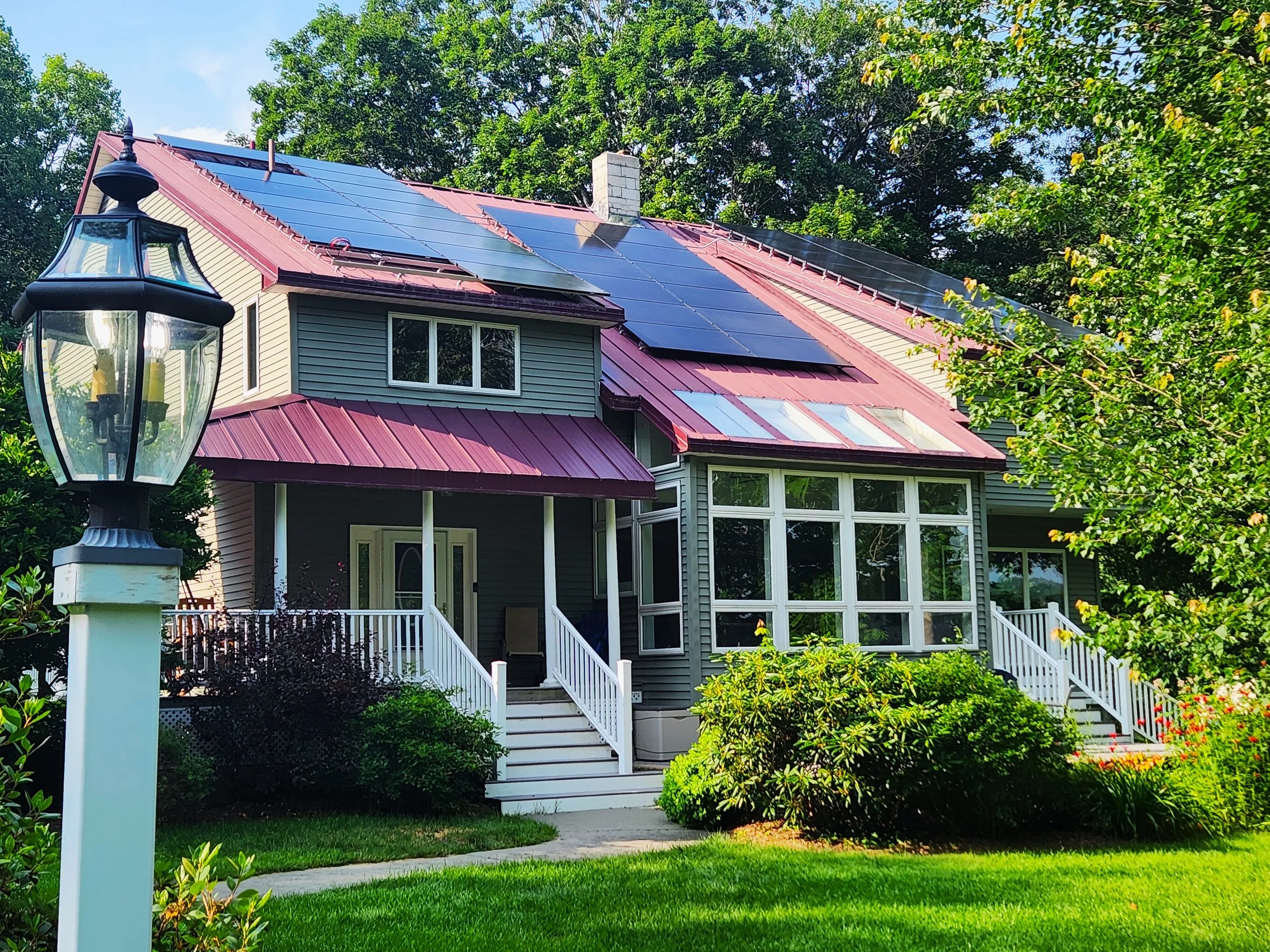 A house with solar panels on the roof, a front porch, and a landscaped yard with bushes, trees, and a grassy area, under a blue sky.
