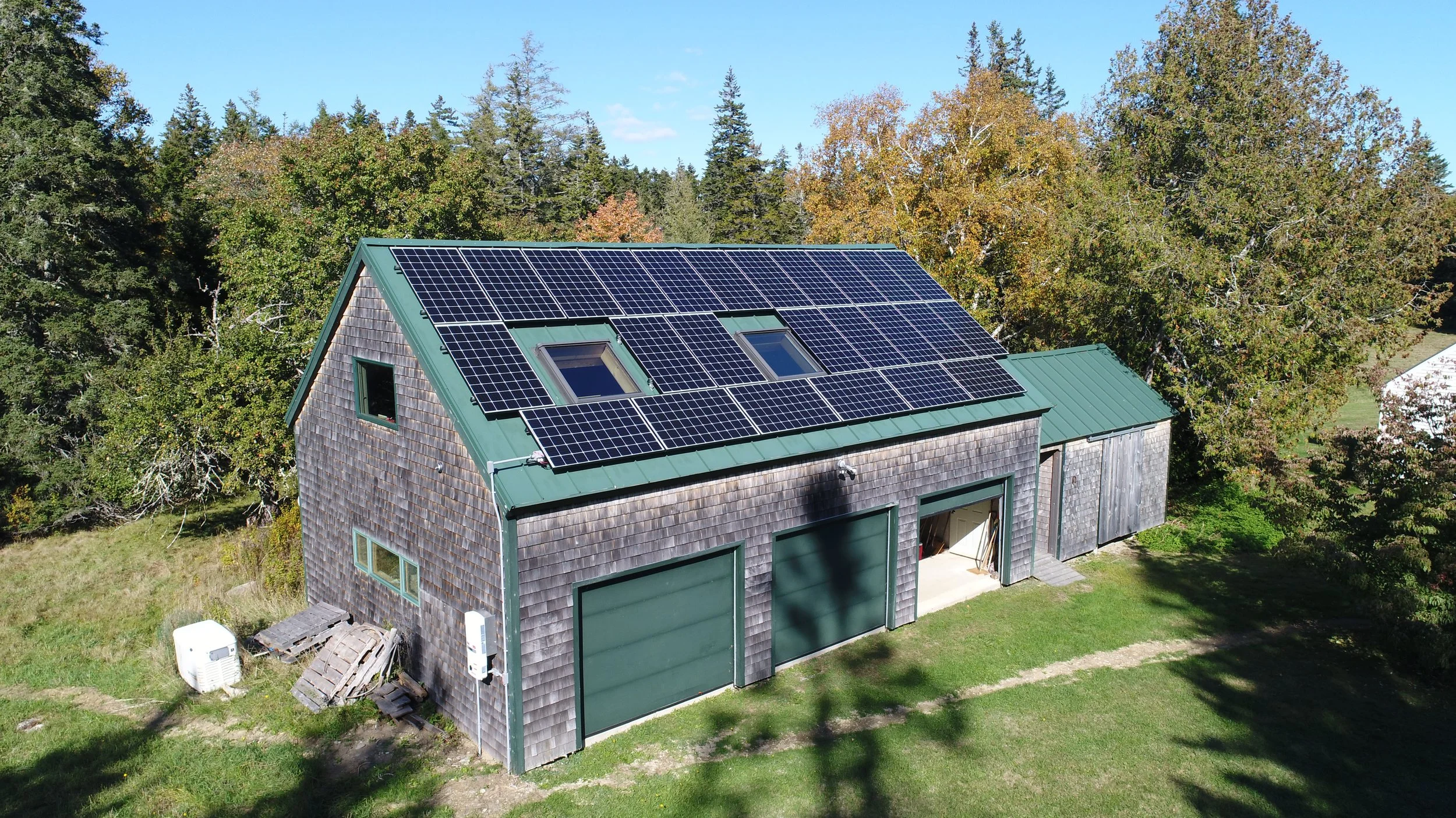 A house with solar panels on its roof, surrounded by trees with autumn foliage, grass, and a blue sky.