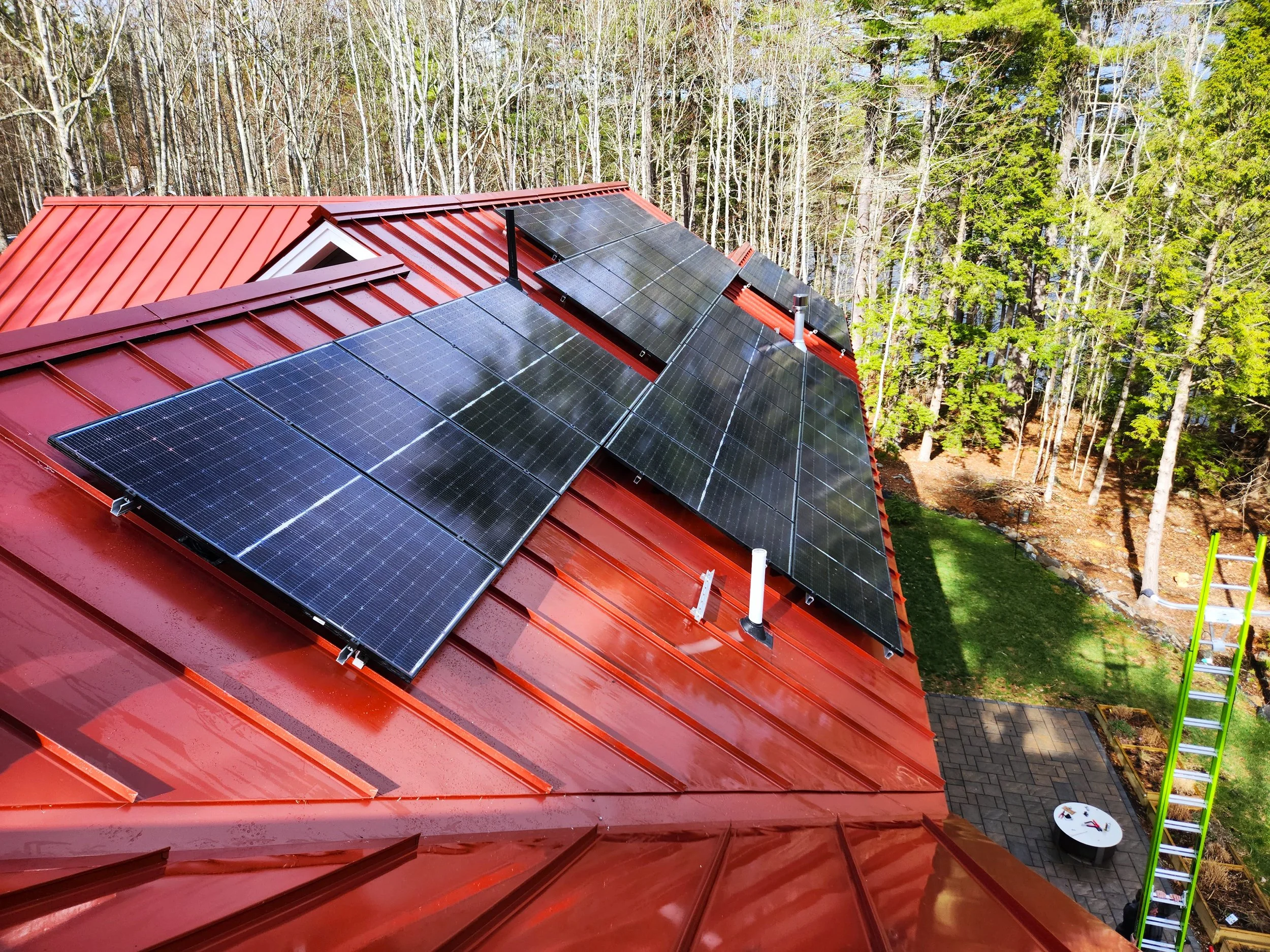 A house rooftop with red metal roofing tiles fitted with solar panels, surrounded by a wooded area with tall trees.