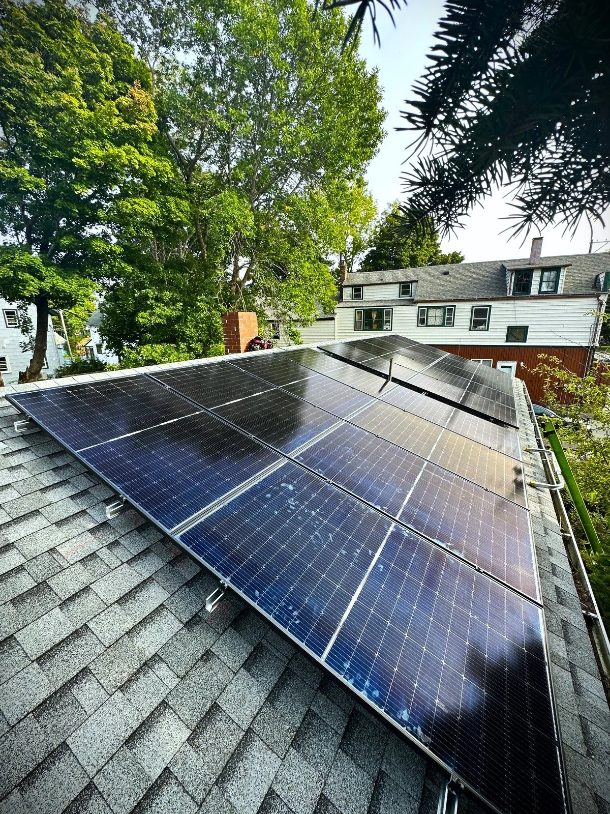 Solar panels installed on a residential roof, with trees and neighboring houses in the background during sunny weather.