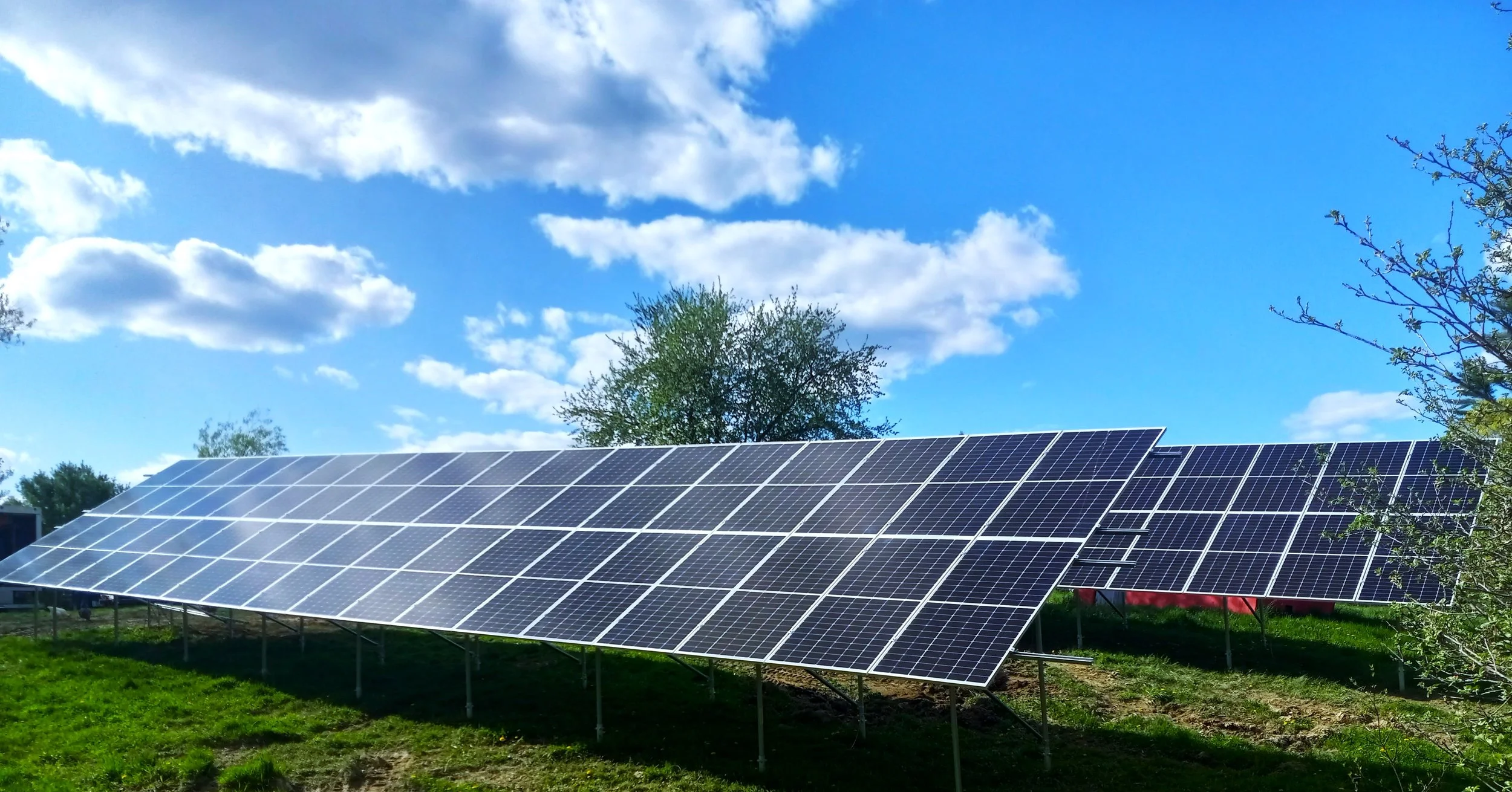 A large array of solar panels installed outdoors on a grassy field, with a background of trees and a partly cloudy blue sky.