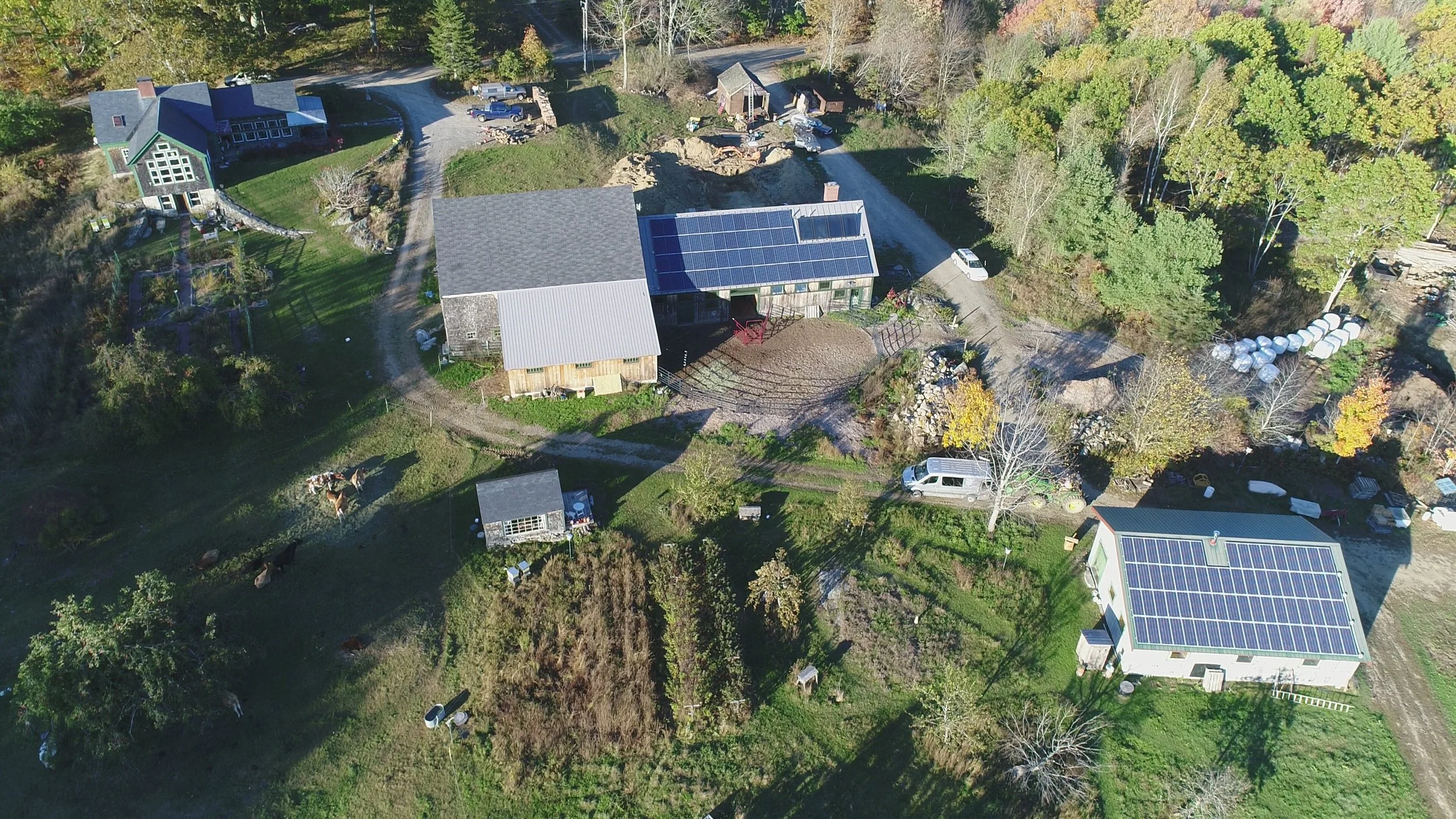 Aerial view of a rural property with houses, some with solar panels, surrounded by trees and grass, with several vehicles and small structures present.