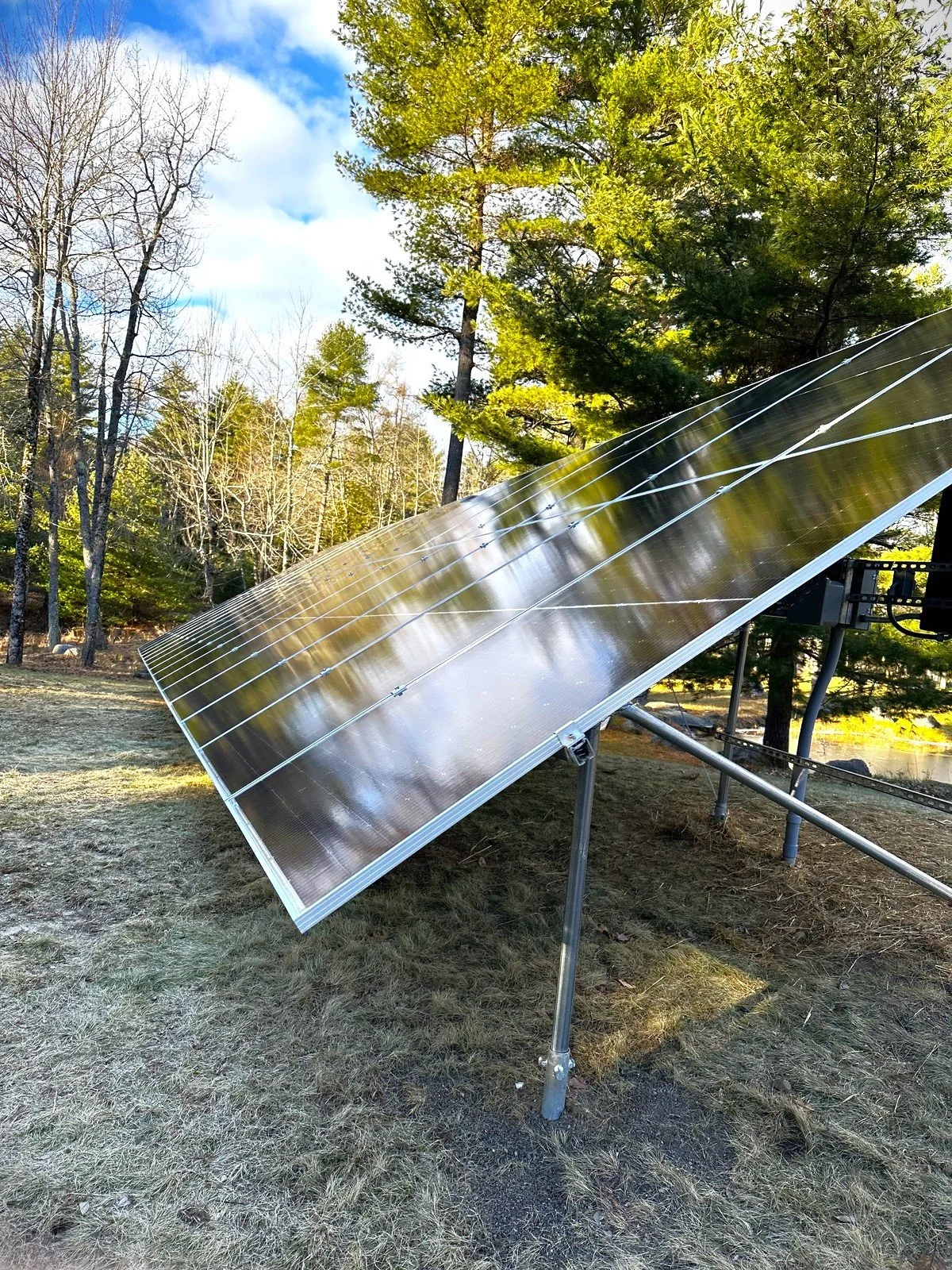 A large solar panel tilted on a metal stand outdoors in a grassy area with trees and a blue sky overhead.
