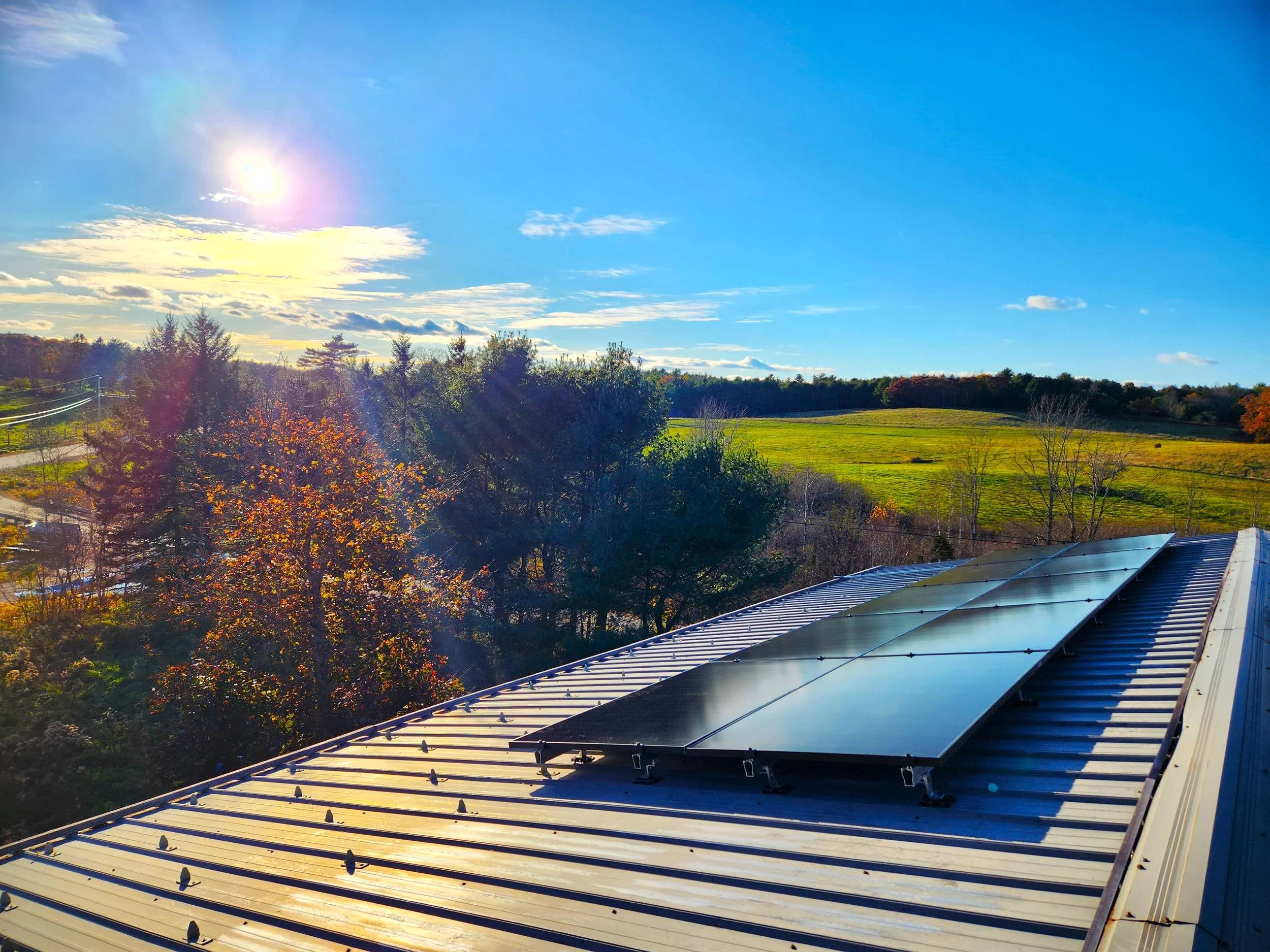 Sunny landscape with green fields, trees with colorful autumn leaves, and a metal roof in the foreground, under a bright blue sky with scattered clouds.