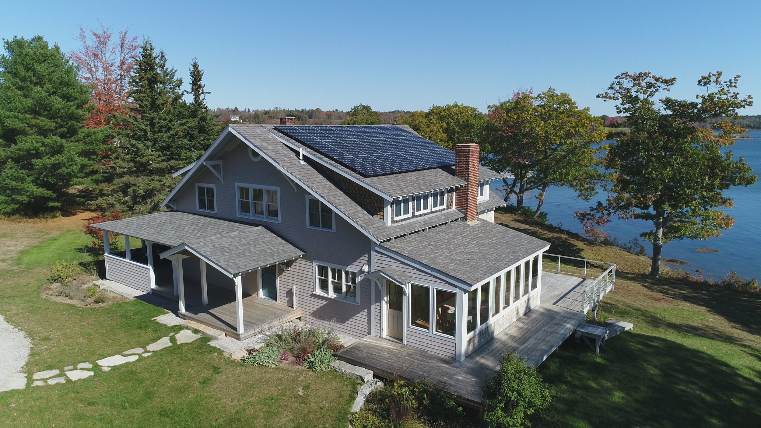 A house near a large body of water with trees around it and solar panels on the roof.