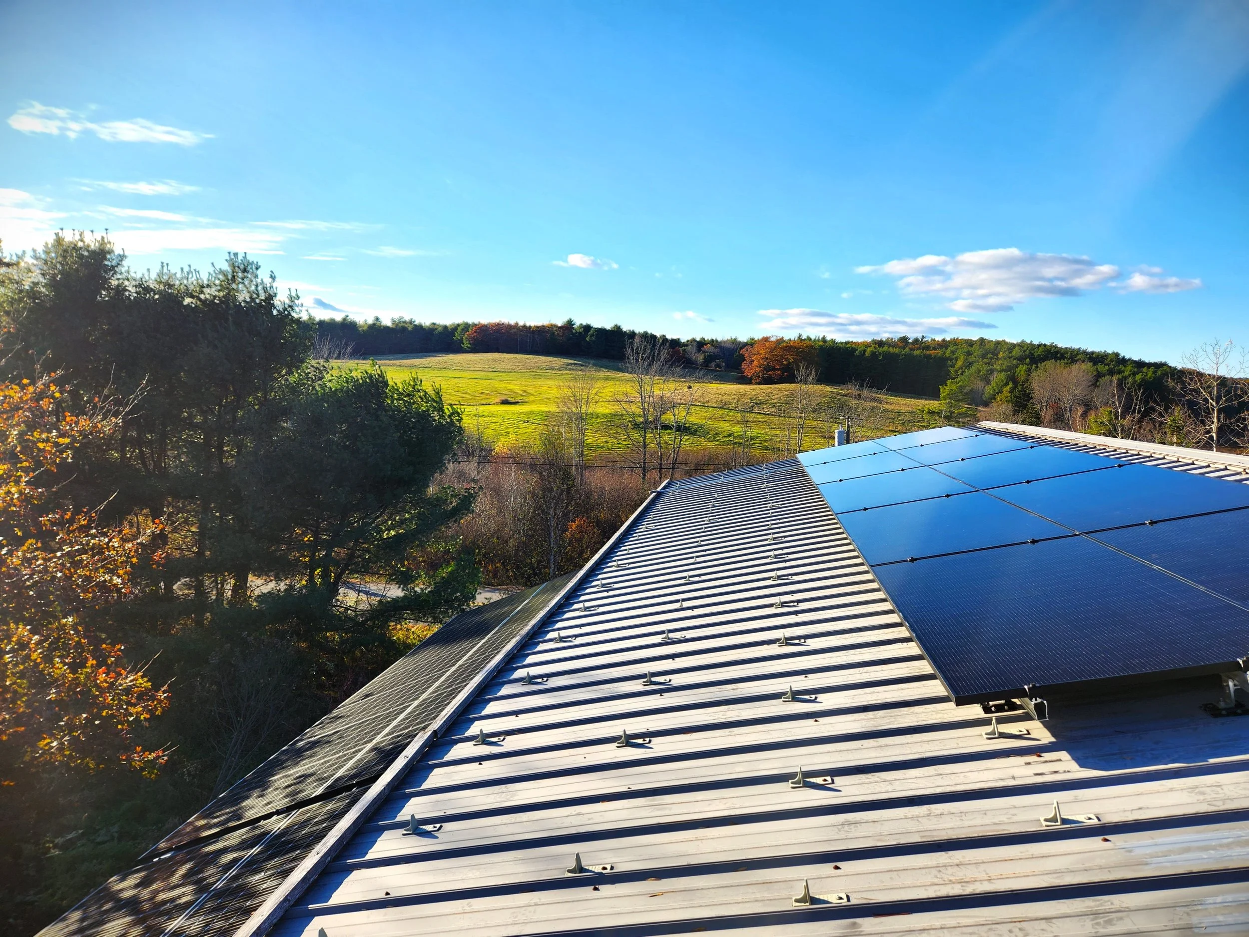 Solar panels installed on a metal roof with a scenic landscape of trees, open fields, and a bright blue sky in the background.