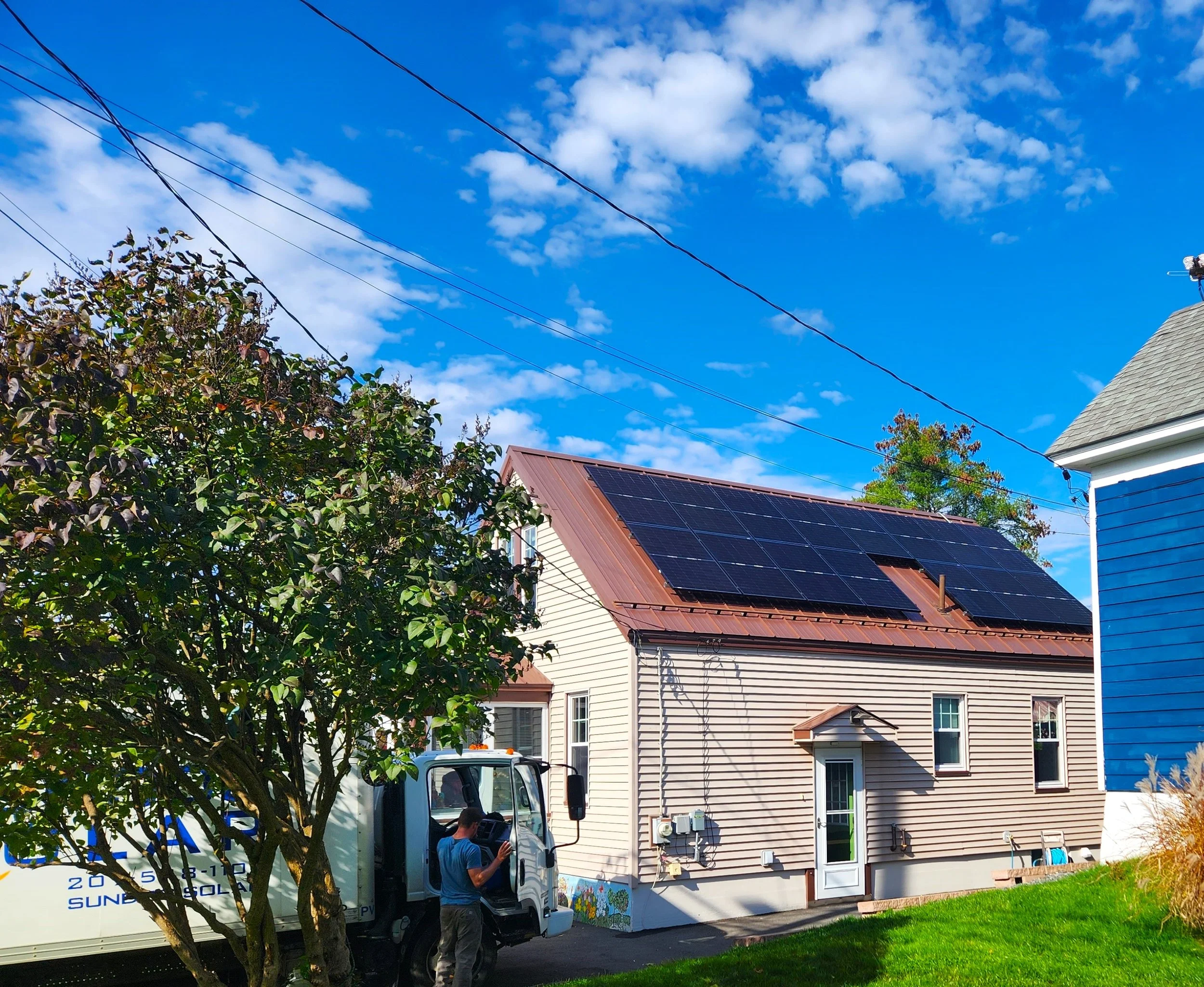 A residential house with solar panels on the roof, a tree in front, and a moving truck and worker on the driveway under a blue sky with scattered clouds.