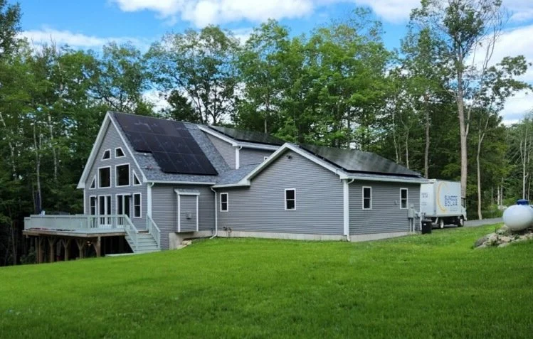 Gray house with solar panels on roof, large windows, and a small deck, surrounded by green yard and trees.