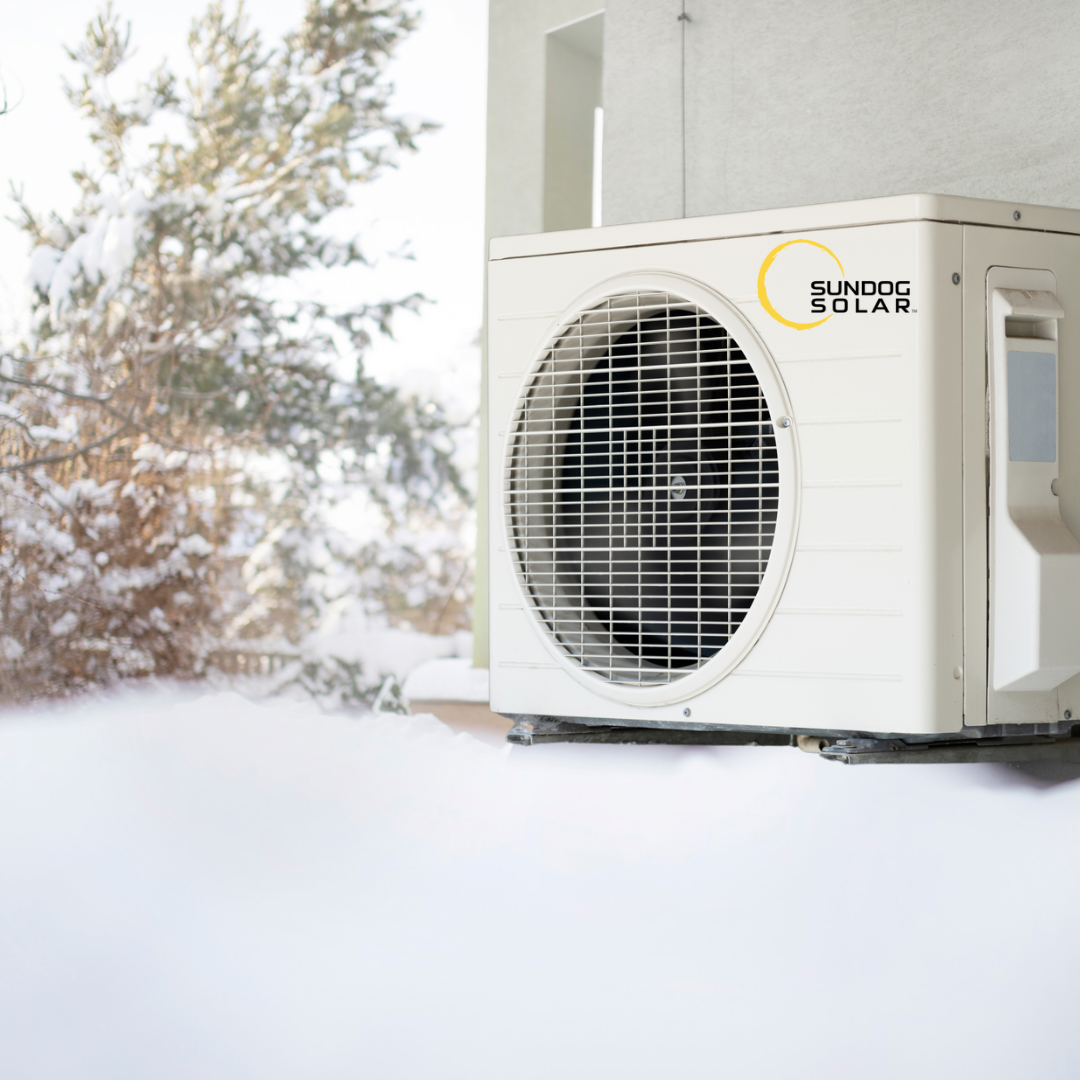 An outdoor solar-powered air conditioning or heat pump unit with the brand name Sundog Solar, situated on a snow-covered surface beside a building. Snow-covered trees are in the background.