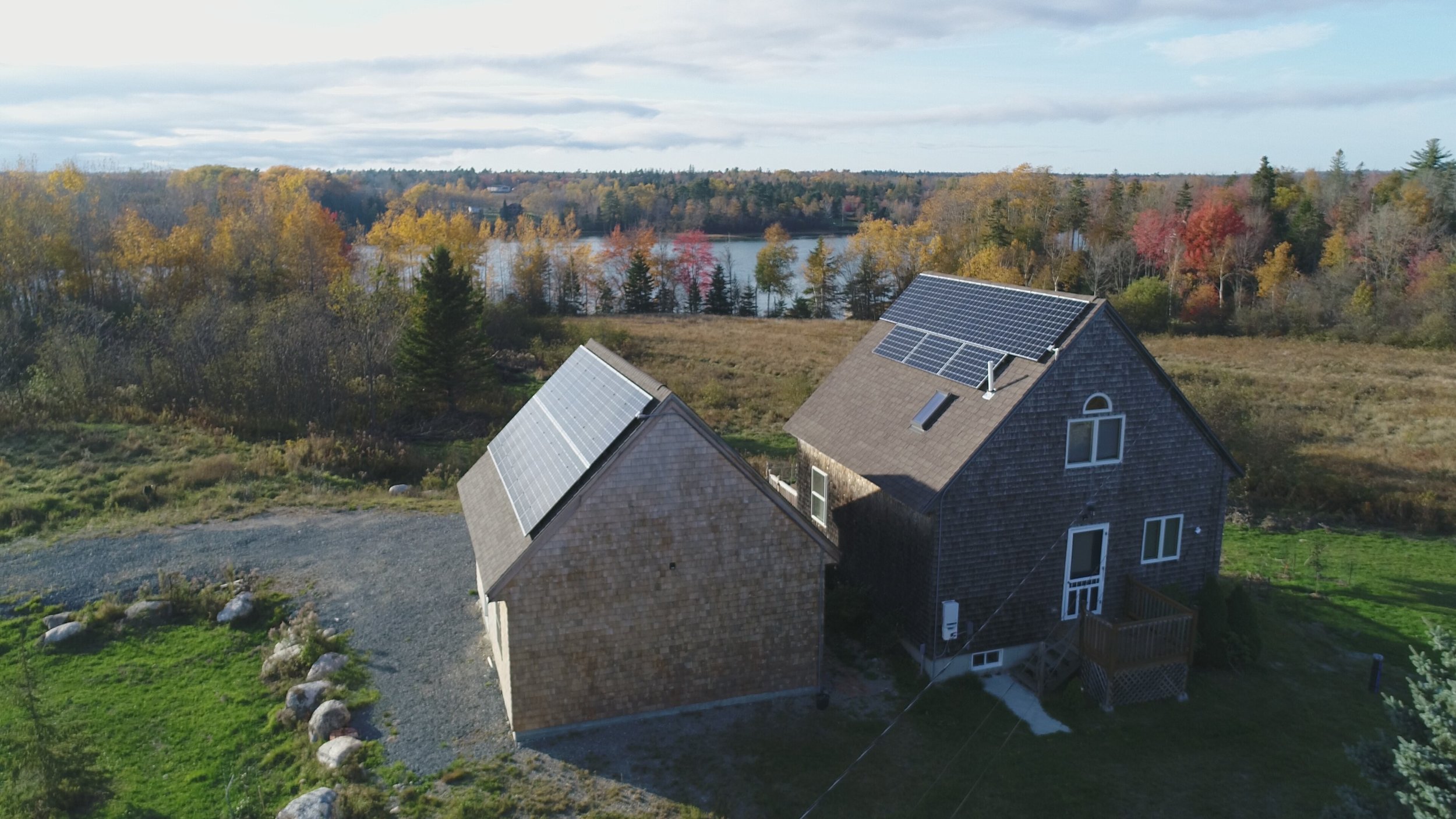 A house with solar panels on the roof, situated in a rural area with trees and a body of water in the background, during autumn.