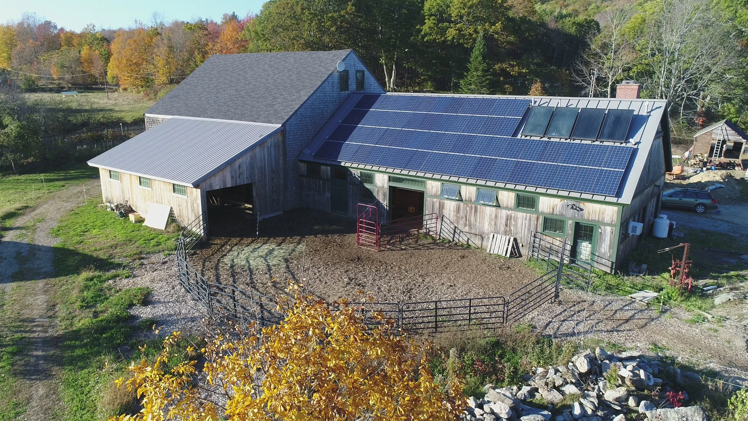 Aerial view of a farm building with solar panels on the roof, surrounded by trees with fall foliage.