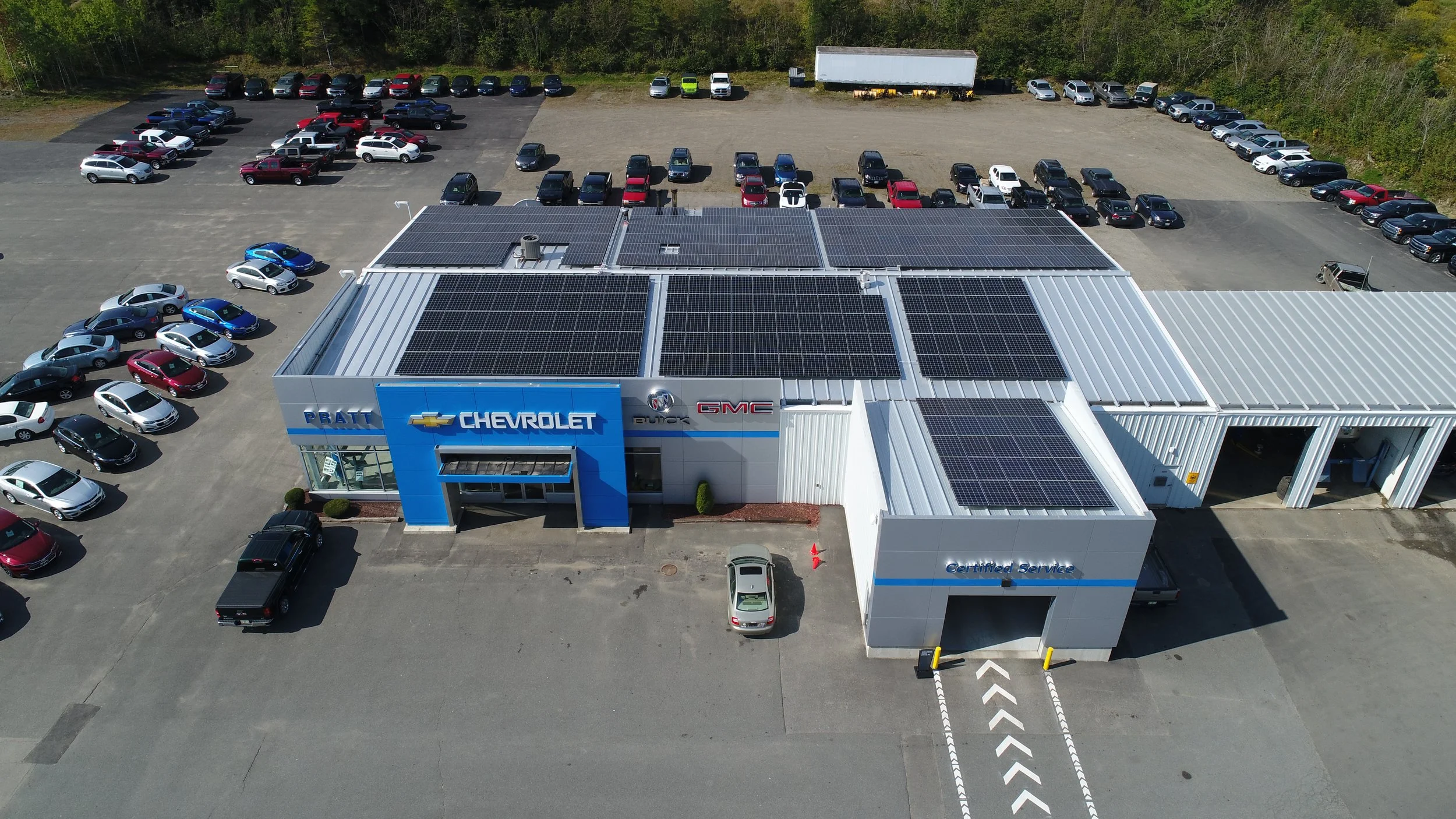 A car dealership building with solar panels on the roof, surrounded by a parking lot full of various vehicles, and a forested area in the background.